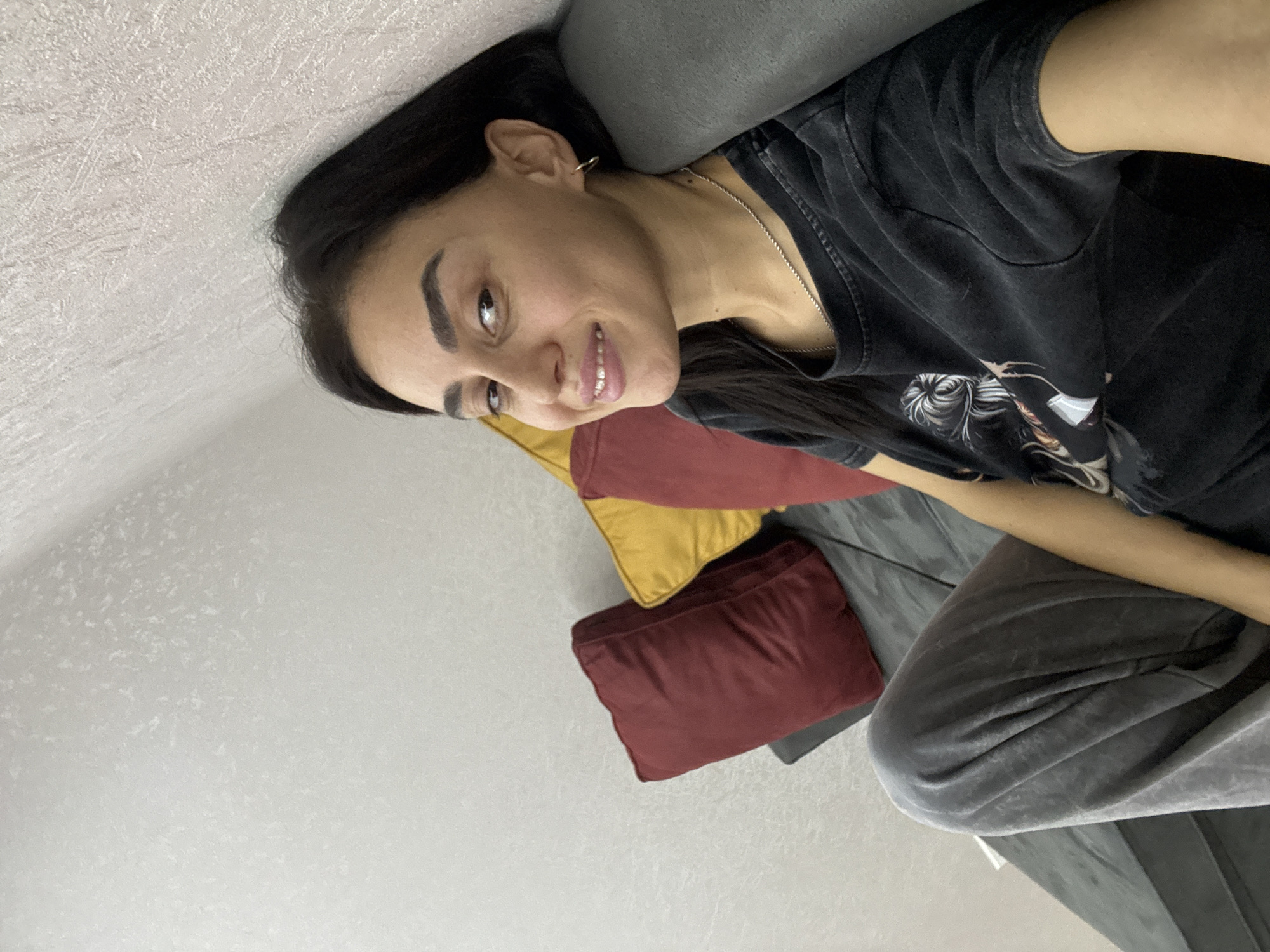 A young woman with dark hair and dark eyes smiles at the camera while leaning against a gray couch. She is wearing a black t-shirt and gray sweatpants. The couch is adorned with two decorative pillows, one red and one yellow. The background is a textured white wall. The scene appears to be an indoor setting, possibly a living room, in Tbilisi, Georgia.