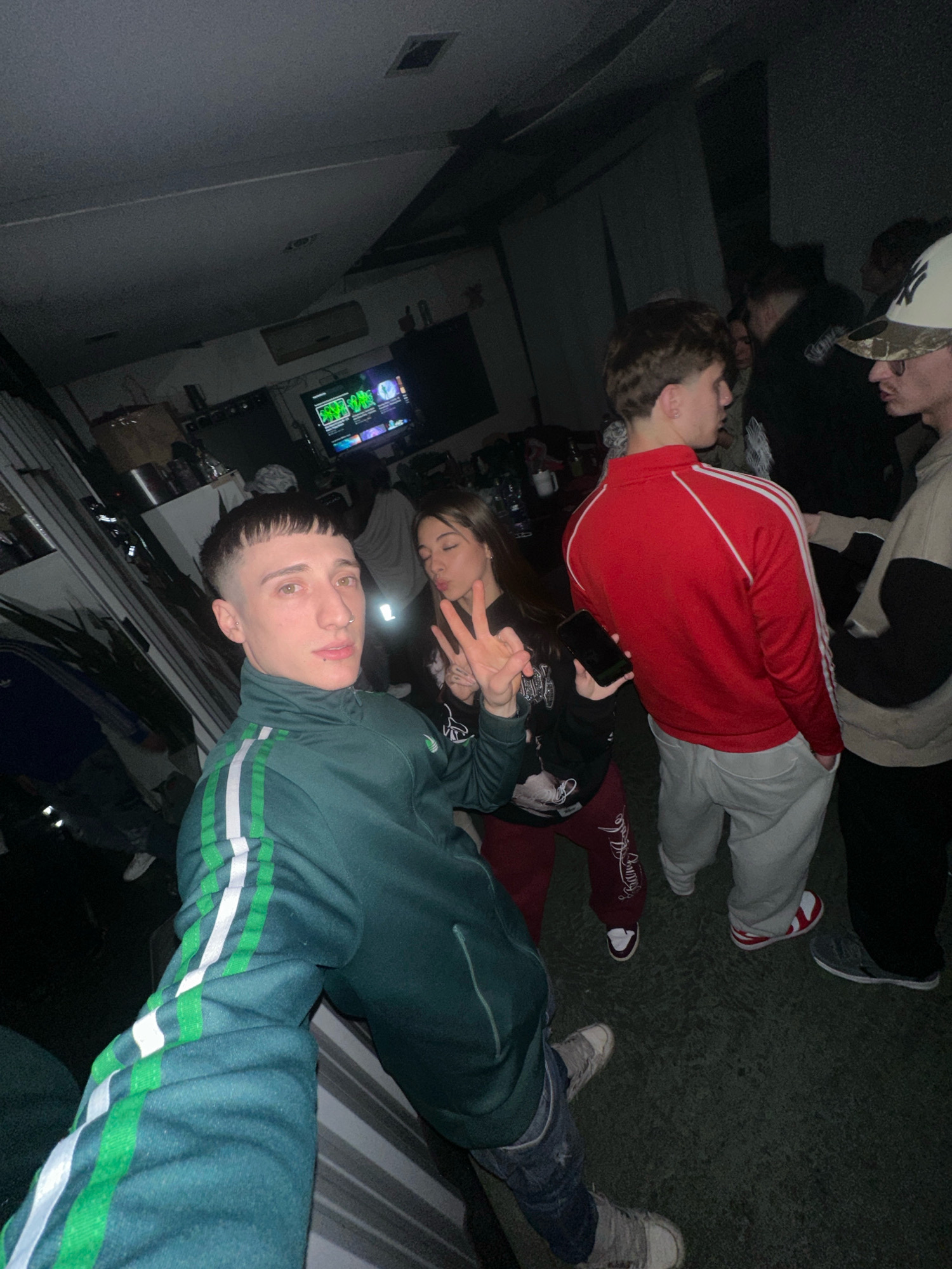 In Valentín Alsina, Argentina, a group of young adults are gathered indoors, possibly at a party. The lighting is dim, with some ambient light sources visible. In the foreground, a person wearing a dark green tracksuit with white and green stripes is looking at the camera and making a peace sign with their fingers. To their right, a young woman is also making a peace sign, and another person in a red jacket and light grey pants is visible in the background. Other individuals and objects like a television are present in the dimly lit room. The floor appears to be a dark, textured surface.