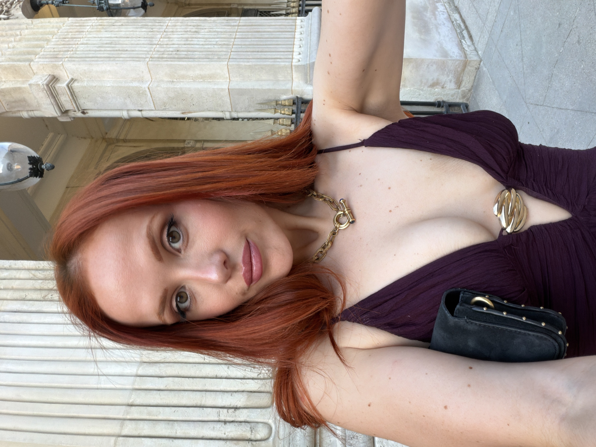 A woman with red hair is shown in a selfie, wearing a dark purple dress and gold jewelry. She holds a small black purse against her chest. The background suggests an architectural structure in Paris, France, featuring light-colored stone with classical detailing, potentially a palace or historical building. A street lamp can be seen near the top left corner of the frame. The woman is looking into the camera, slightly tilted to her left.