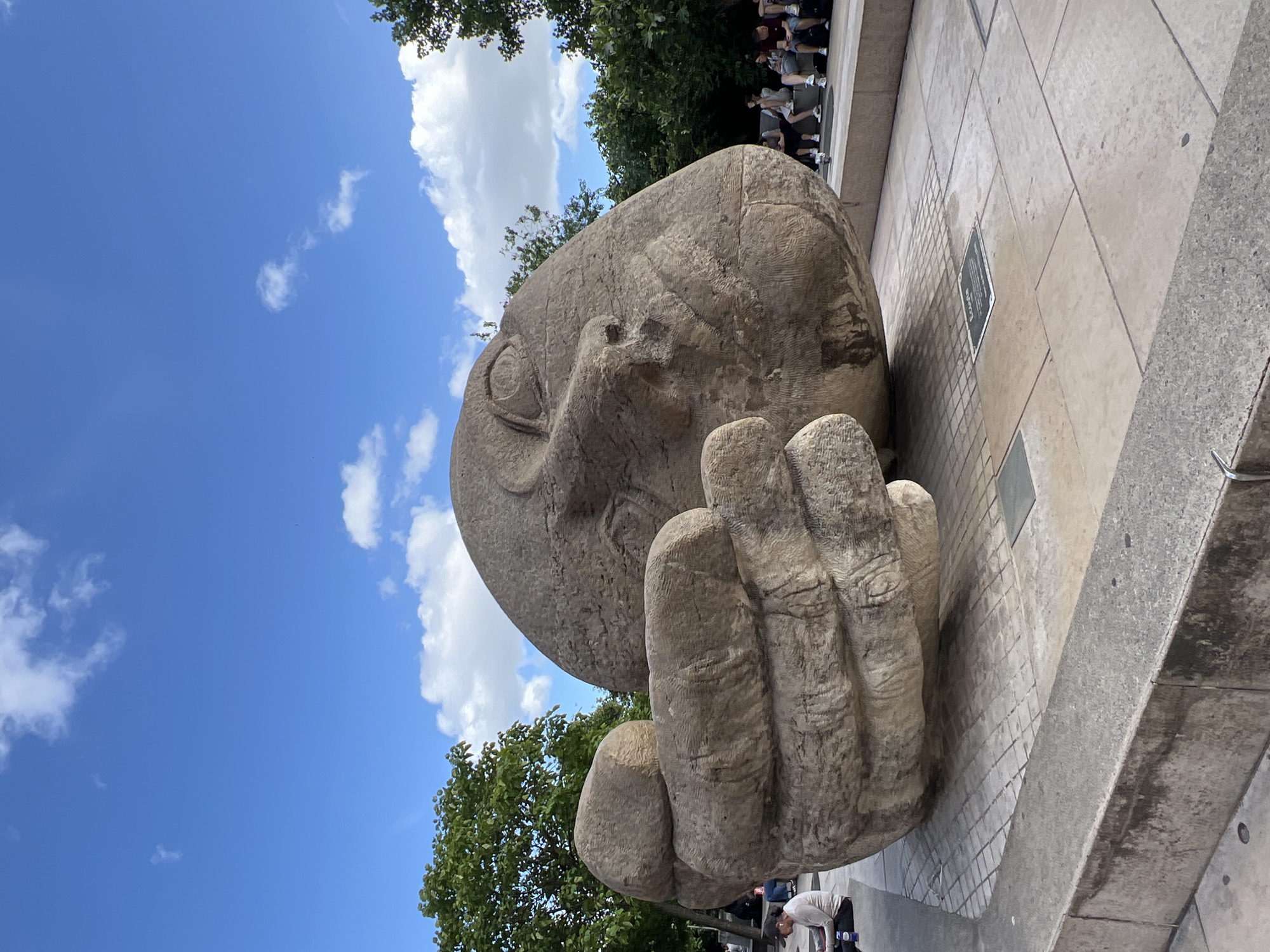 The image shows a large, carved stone sculpture of a disembodied head and hand lying on a paved plaza. The sculpture is weathered and has visible tool marks. The head is resting on its side, with its features sculpted in a stylized manner. A large fist is positioned in front of the head. The sculpture is situated outdoors, with a clear blue sky with scattered white clouds overhead. Lush green trees are visible in the background. Several people are seated or standing around the plaza, some interacting with the sculpture or with each other. One person is sitting on the ground to the left of the sculpture, looking down at something. Another group of people is seated on a bench in the background to the right. A metal plaque is embedded in the paving stones in front of the sculpture.
This sculpture is known as "Le Monument à la Tête qui pense" or "The Monument to the Thinking Head" and is located in the Jardin des Tuileries in Paris, France.
