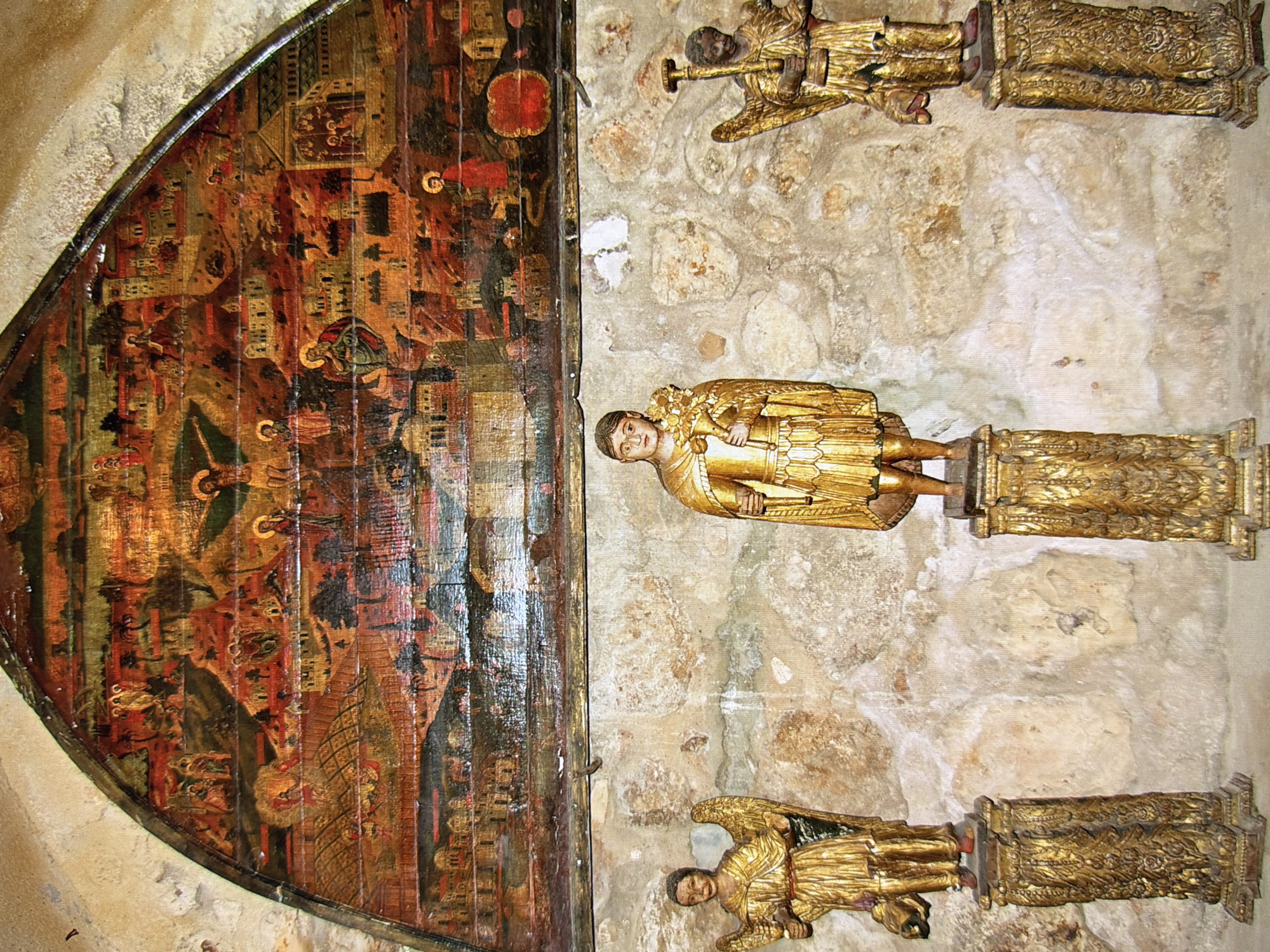The image shows a religious fresco painted on a wooden panel above three golden statues, likely saints or angels. The fresco depicts a biblical scene with multiple figures, buildings, and landscapes rendered in warm, earthy tones. The three statues stand on ornate golden pedestals against a textured stone wall. The statue on the left appears to be an angel with wings, holding an object in its left hand. The central statue is of a male figure holding a bouquet of flowers. The statue on the right is also an angel, holding a staff-like object. The overall scene evokes a sense of historical and religious significance. This fresco and statues are located in the St. Nicholas Roman Catholic Cathedral in Kyiv, Ukraine.