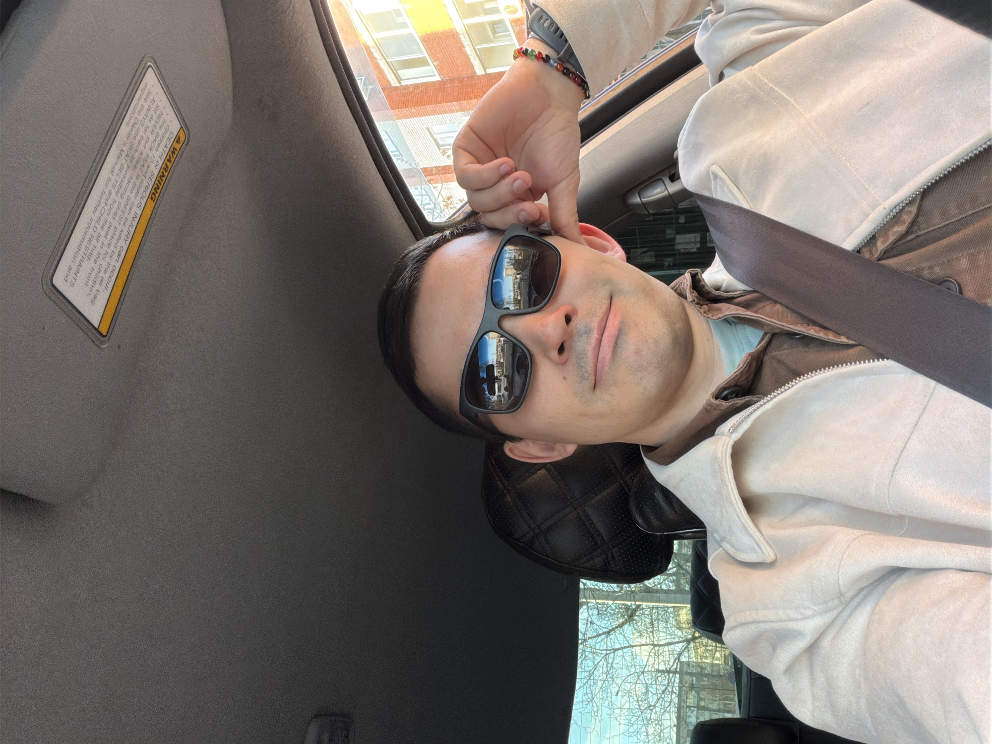 A person is seen from a slightly upward angle, likely taking a selfie inside a car during the daytime. The individual, who appears to be male, wears dark sunglasses, a light beige jacket over a brown inner layer and a light blue top, and a gray seatbelt across their chest. A multi-colored beaded bracelet is visible on their right wrist, and their right hand is positioned near their head. The car's interior includes a gray headliner and a sun visor with a prominent warning label. Through the car windows, brick buildings, a clear sky, and bare tree branches suggest a colder season. The visible text on the sun visor's warning label reads: "A WARNING DEATH or SERIOUS INJURY can occur. The BACK SEAT is the SAFEST place for children. Children 12 and under can be killed by the air bag. NEVER put a rear-facing child seat in the front. ALWAYS use SEAT BELTS and CHILD RESTRAINTS. Sit as far back as possible from the air bag. See the Owner's Manual for further information and explanations." The person's expression is neutral, partially obscured by the sunglasses. The setting is Izhevsk, Russia.