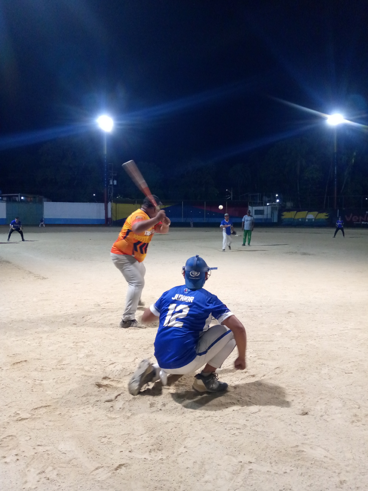 This image captures an active baseball or softball game taking place at night on a dirt field. The perspective is from behind home plate, looking towards the pitcher and outfield.
In the immediate foreground, a catcher, wearing a blue helmet and a blue jersey with "JUNIOR" and the number "12" emblazoned on the back, crouches in position. Directly in front of him, a batter in an orange and black jersey and light grey pants is in mid-swing, holding a baseball bat. His back is towards the viewer, indicating a powerful swing or follow-through.
The field is brightly illuminated by multiple powerful floodlights, which are visible as intensely bright points in the dark night sky, creating distinct lens flares. In the mid-ground, a pitcher wearing a white shirt and blue pants can be seen, with a small white ball suspended in the air nearby, suggesting a pitch has just been delivered or hit. Another player in a green shirt and dark pants stands further back, observing the play. More distant players are faintly visible across the field.
The background features a fence or backstop, adorned with banners. One banner prominently displays the Venezuelan flag, while another banner further to the right includes the partially visible text "Movilnet". Trees line the very far background, silhouetted against the night sky. The scene conveys the focused energy of a competitive sports event under artificial lighting.