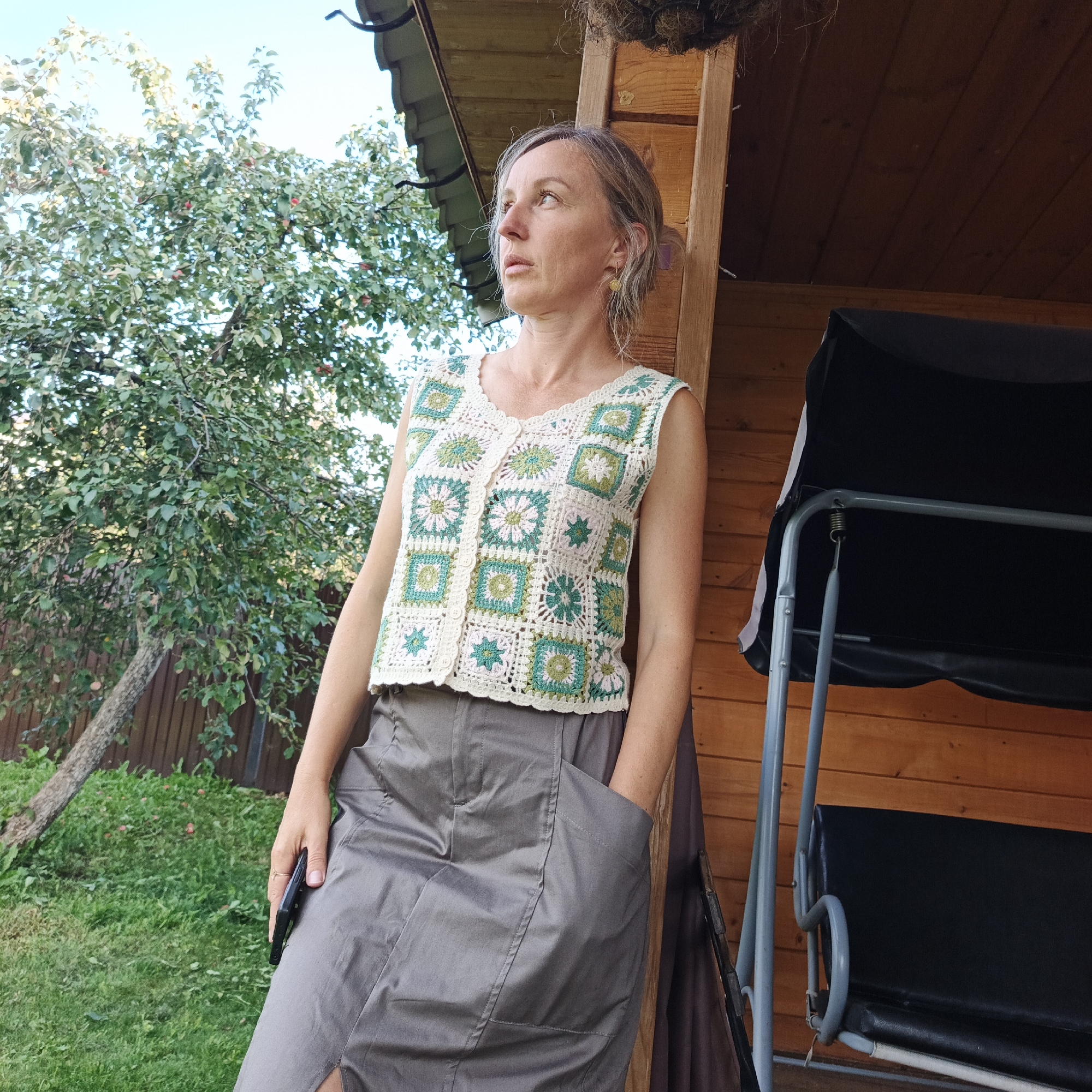 A woman with light brown hair pulled back stands leaning against a wooden post next to a wooden structure. She is wearing a white crochet vest with green and pink floral square patterns and a gray skirt. Her right hand is tucked into a pocket of her skirt, and her left hand holds a black smartphone. In the background, there is a green tree with some red fruit, a wooden fence, and a portion of a black swing set. The scene is outdoors during the daytime. The woman is looking off to the left.