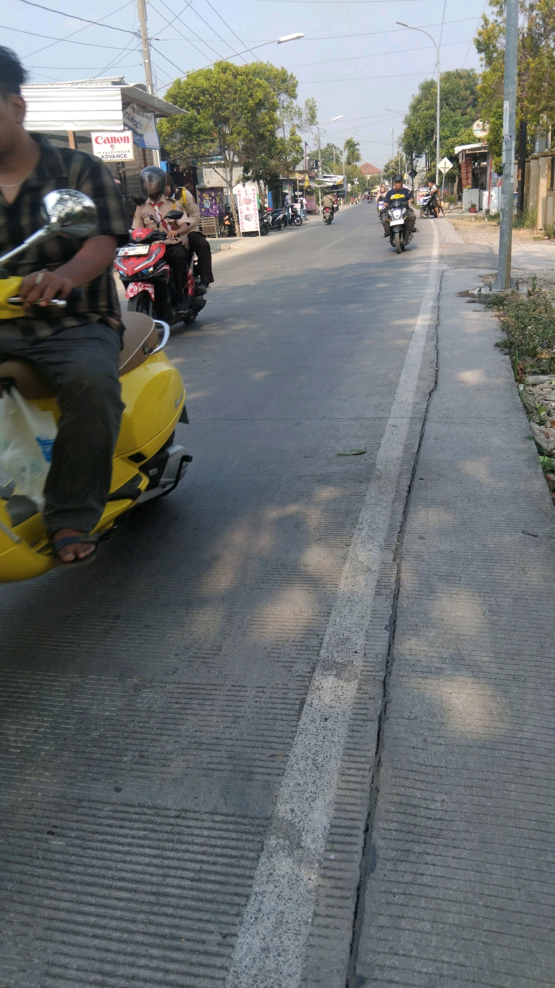 The image depicts a street scene in Tegal, Indonesia, with several motorcycles and riders. In the foreground, a person is riding a yellow scooter, facing away from the camera. The rider is wearing a dark plaid shirt and dark pants. To the left of the scooter, there is a white bag. On the road, a red motorcycle with two riders is moving away from the camera; the rider in front wears a brown uniform with a red scarf, and the person behind them is wearing a dark uniform and a helmet. Further down the road, another person is riding a black motorcycle. The street is paved and has a white line marking the edge of the road. Along the side of the road, there are buildings, trees, and some vegetation. The sky is clear and bright.