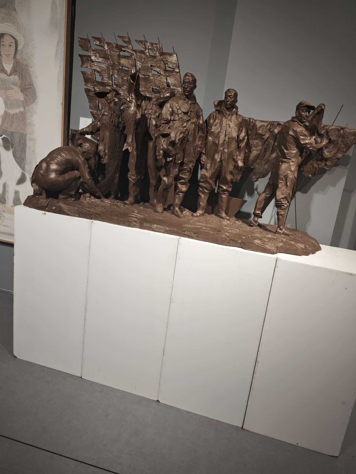 The image shows a bronze sculpture depicting a group of figures, presumably engaged in some form of labor. The sculpture is placed on a white rectangular base, constructed from several vertical panels.
To the left, a painting hangs on the wall showing a woman and a dog. The background wall is a neutral gray color. The setting appears to be an art gallery or museum, with the sculpture on display. The location is Unknown, Unknown.