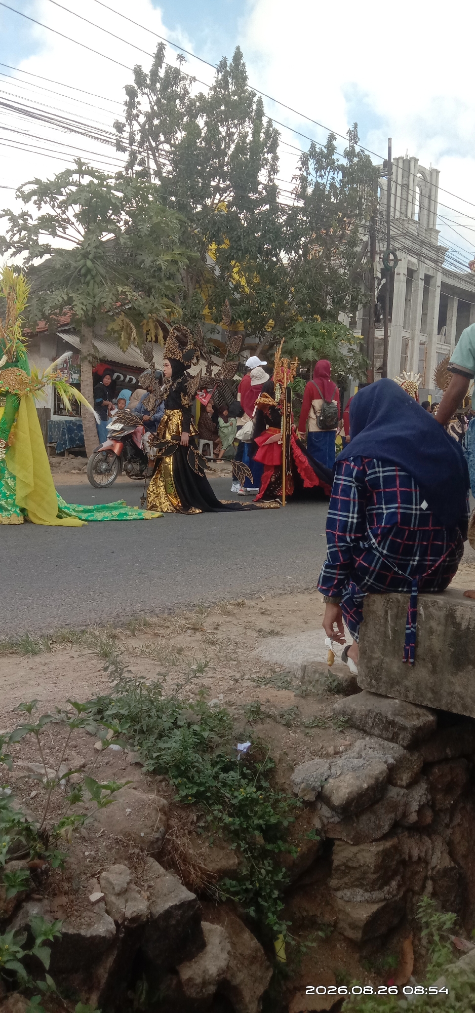 In Pakong, Indonesia, a cultural parade is underway on a paved road. The foreground shows a person in a blue and white plaid hijab and shirt, sitting on a concrete structure. They are holding a white object, possibly a tissue. Behind them, several individuals are dressed in elaborate traditional costumes. One prominent figure wears a black and gold ornate dress with intricate headwear and wing-like embellishments. To the left, another person is adorned in a bright green and yellow costume with feathered headpieces. Several other participants in similarly vibrant attire are visible further down the road. Spectators, including children and adults, are gathered on the side of the road, some near a parked motorcycle and a modest building. Trees with green foliage line the street, and in the background, a large, classical-style building with arches and columns stands against a cloudy sky. Power lines are visible overhead. The overall scene depicts a community celebration with a focus on traditional dress and performance.