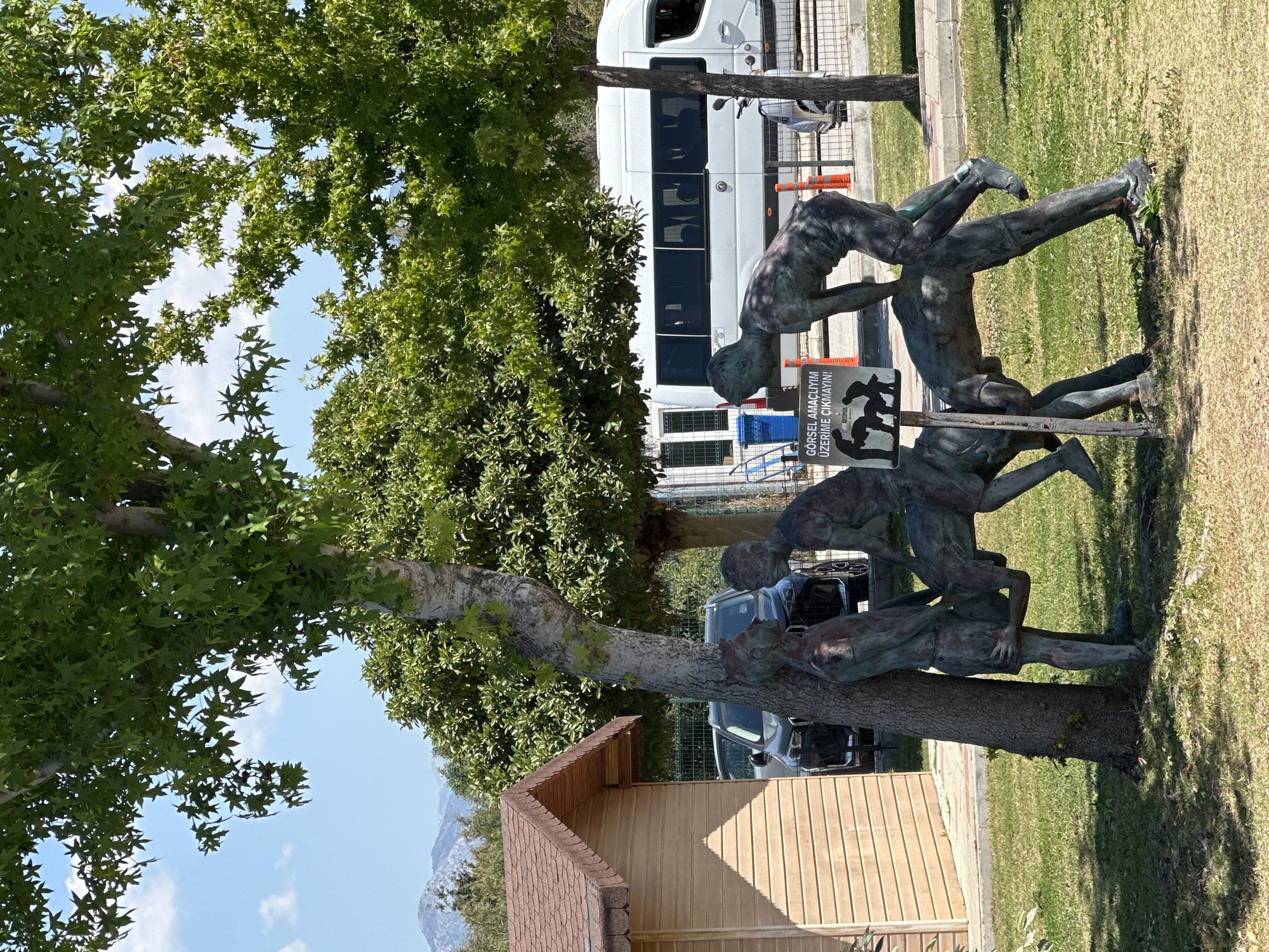 The image shows a sculpture in a park setting. The sculpture appears to depict a group of people climbing or being supported by each other, made of a dark, possibly bronze-like material. A sign next to the sculpture has text in Turkish. The sculpture is located near a tree. In the background, there is a white van, a building with a brown roof and wooden walls, possibly a shed or small structure, and other greenery. The sky is visible through the trees. Based on the location, it can be inferred that this might be in Fethiye, Turkey.