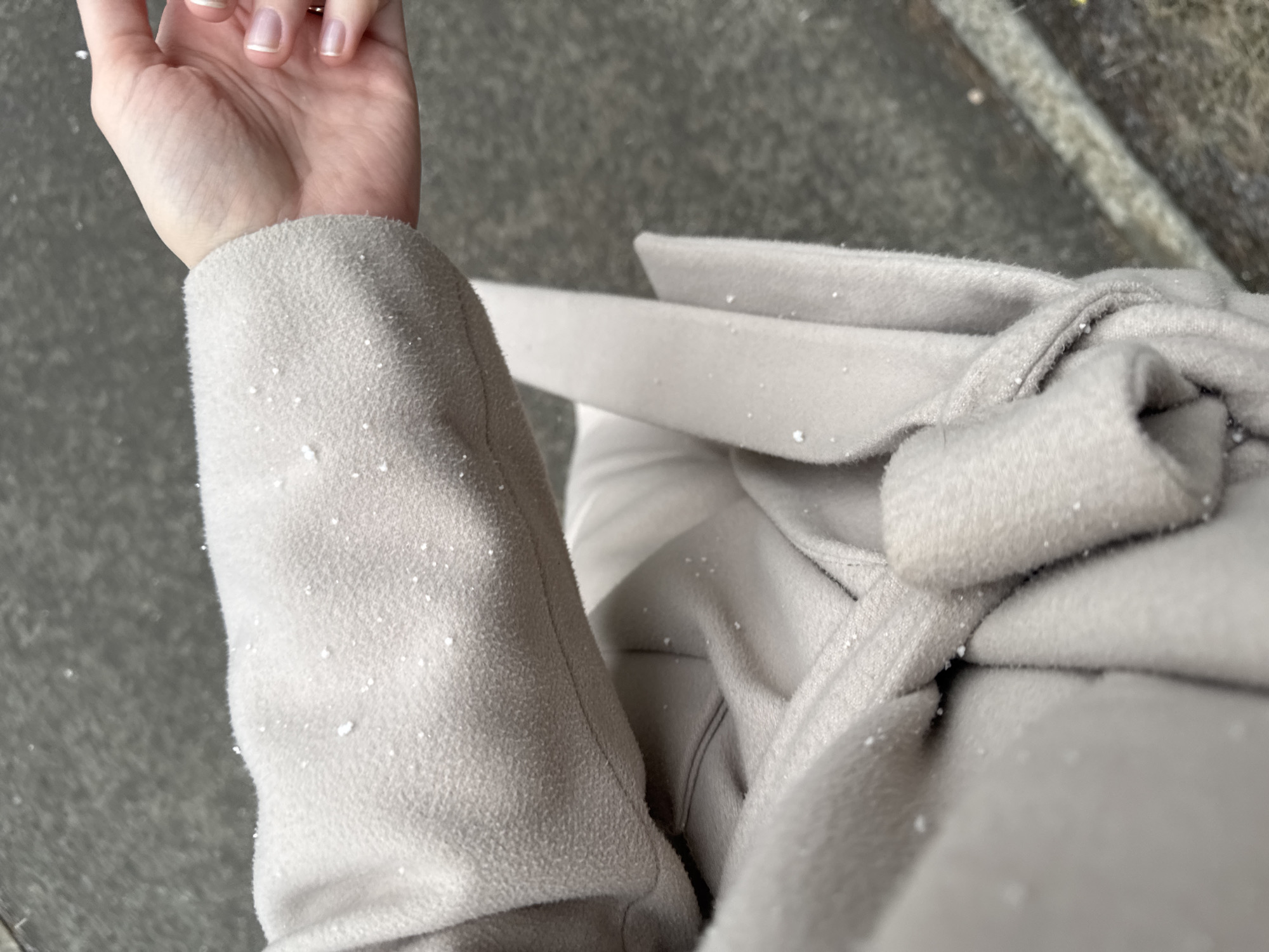 An overhead shot shows a person's left arm and the front of their beige wool coat covered in snowflakes. The person's hand, with manicured nails, is visible at the top of the frame. The coat is tied with a belt. The background shows a gray concrete surface with a slight edge of a stone curb. Given the details, the location is likely Perm, Russia, due to the presence of snow and associated winter clothing.