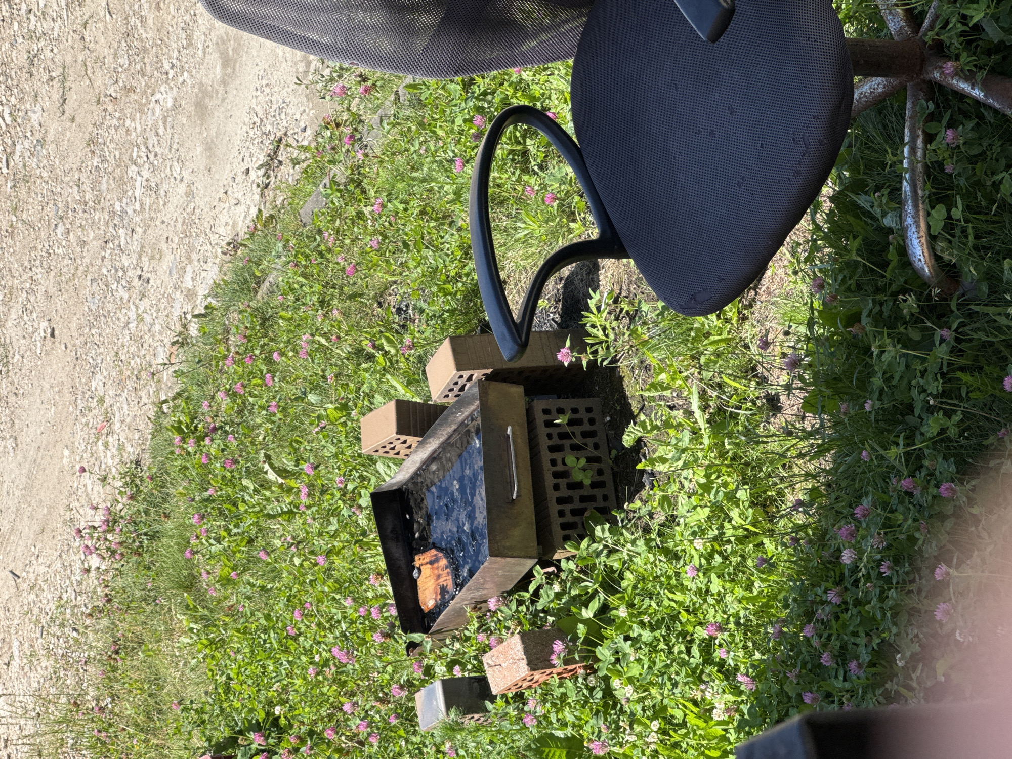 In a grassy area with scattered pink clover flowers, a portable metal grill is set up on top of stacked bricks. The grill contains charcoal and a piece of wood. To the left and right of the grill are additional bricks, one of which is a lighter color with holes. An office chair with a dark fabric seat and backrest is positioned to the right of the grill. The ground behind the grassy area is a rough, gravelly surface. The scene appears to be outdoors in a rural or semi-rural setting, possibly on the outskirts of the city of Zaokskiy, Russia, given the context provided. There are no people visible in the image.