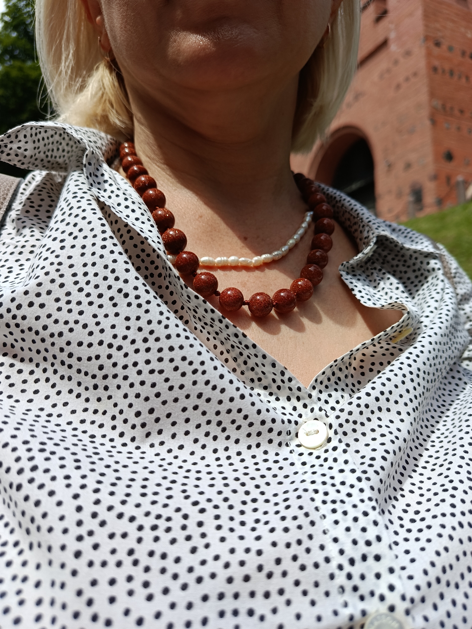 The image shows a close-up of a woman wearing a white shirt with black polka dots and two necklaces. The necklaces are made of brown beads and white pearls. The woman has blonde hair and is outdoors, with a brick building and greenery in the background. The location is not identifiable as Kyiv, Ukraine.