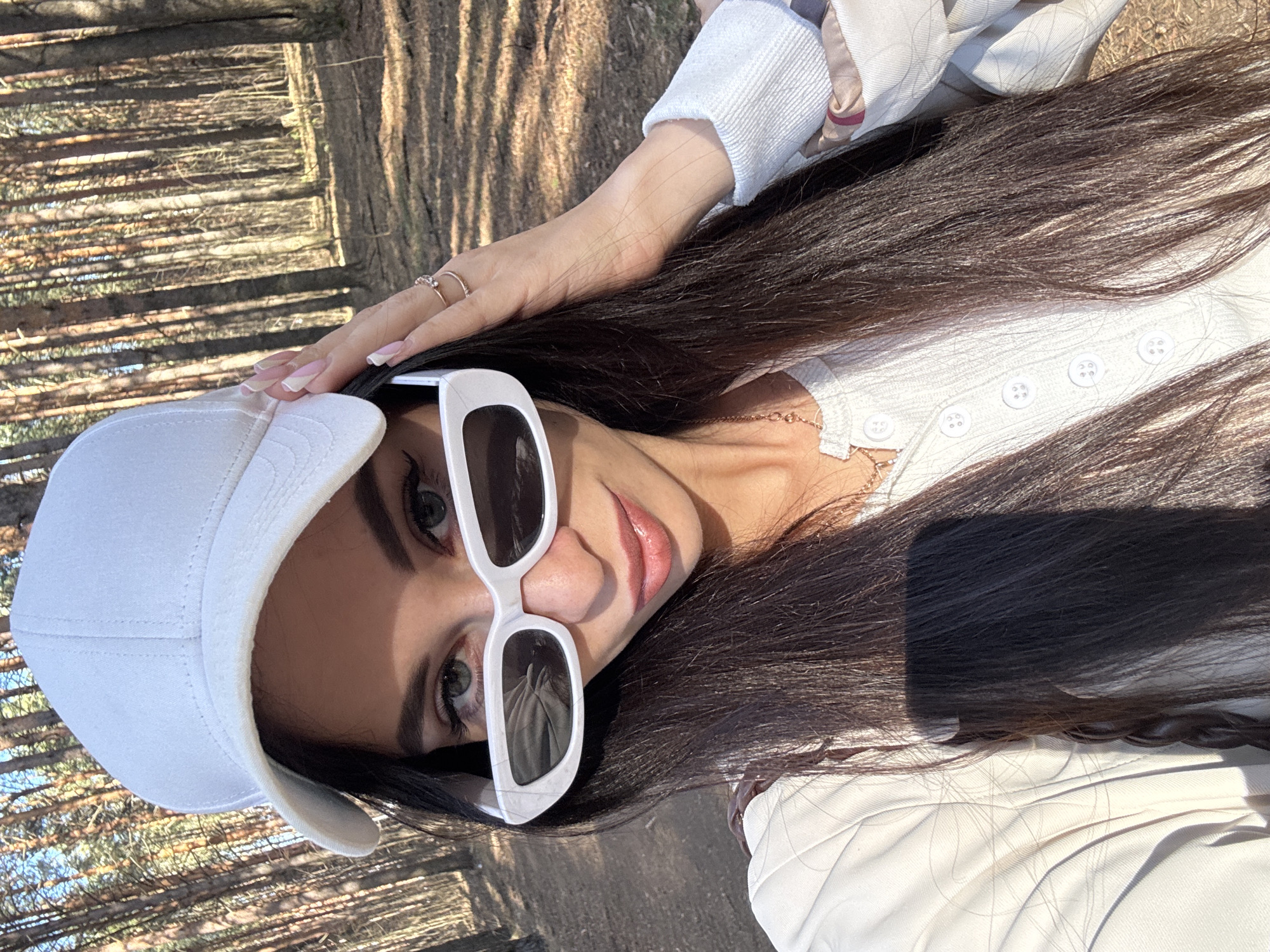A young woman with long, dark hair is photographed outdoors. She is wearing a white baseball cap and large, white-framed sunglasses. Her top appears to be white with buttons. She is wearing rings on her fingers. The background appears to be a forest with sunlight filtering through the trees. The location is Chaikovsky, Russia.