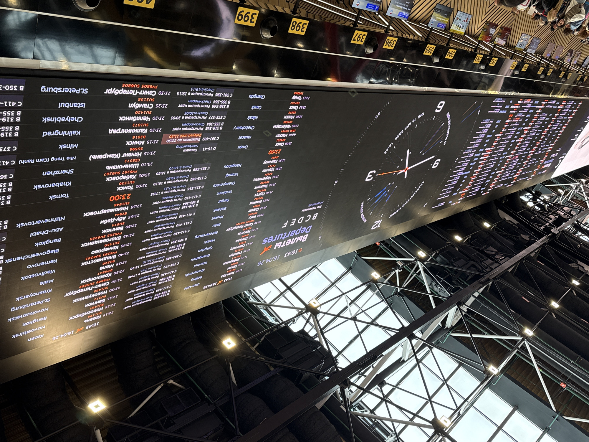 This image captures a wide-angle, tilted view of a large digital departures board within an airport terminal, likely Sheremetyevo Airport in Khimki, Russia. The board, which dominates the scene, displays extensive flight information in both Russian and English, characteristic of an international airport.
The primary subject is the glowing black departure screen, which lists numerous destinations such as St. Petersburg, Istanbul, Chelyabinsk, Kaliningrad, Minsk, Tomsk, Shenzhen, Bangkok, Abu-Dhabi, and many more. Each entry includes flight times (ranging from late evening 21:00s to late night 23:00s), flight numbers (e.g., SU1434, FV6805), and status updates like "Регистрация идет" (Check-in open), "Delayed until 22:50," and "Nowboarding," along with associated gate or check-in counter numbers (e.g., B 355-364, C 411-430).
A prominent digital clock face is integrated into the right side of the main display, showing "THU 16 APR 2026" and the current time "16:43." It also clearly states "MOSCOW RUSSIA" and "SHEREMETYEVO AIRPORT," confirming the location. Above the main screen, a row of yellow-numbered indicators, including "394," "395," "396," "397," "398," and "399," likely denote gate numbers or check-in counters.
The setting reveals modern airport architecture with visible structural beams, overhead lighting fixtures, and a glass ceiling allowing natural light, reflecting on the surface. A few individuals, possibly passengers, are partially visible in the upper right, observing the board or moving through the terminal. The overall activity depicted is passengers checking flight schedules and statuses, a common scene in an active departures hall.