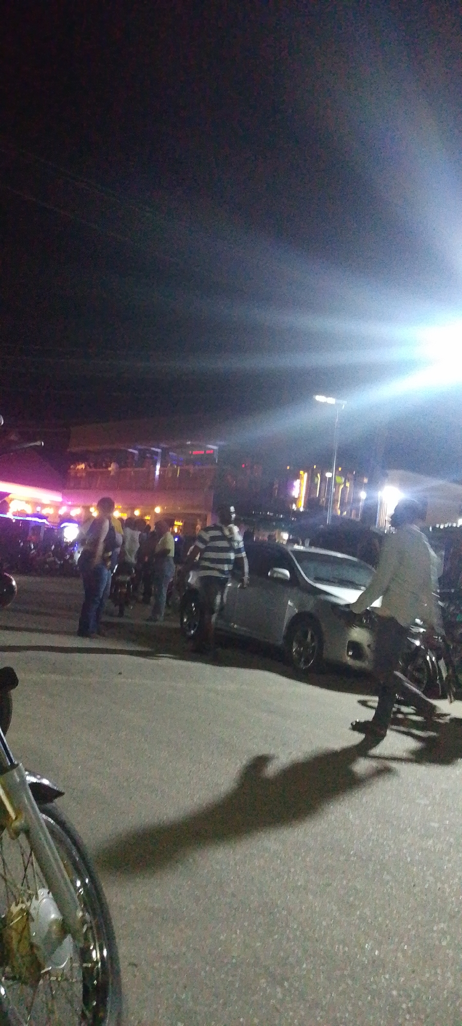 The image shows a street scene at night in what appears to be a commercial or entertainment area. Several people are gathered near a silver car parked on the side of the road. The lighting is poor, with a bright light source creating glare. Some of the people are standing near motorcycles. In the background, a two-story building with lights and what appears to be a second-story balcony can be seen. Based on the prompt this image could potentially be located in the city of Lokoja, Nigeria.
