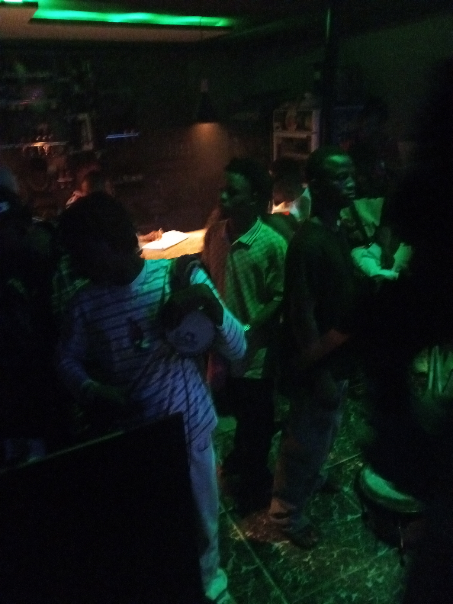 The image depicts a dimly lit indoor space in Lokoja, Nigeria, possibly a bar or club. There are several people present, mostly young men. One person in the foreground wears a blue and white striped shirt. Another man stands next to him wearing a green shirt with a patterned design. Other individuals are partially visible in the background. The lighting is predominantly green, with some light sources visible in the background. The floor has a green and white speckled pattern. A counter or bar is present in the background.