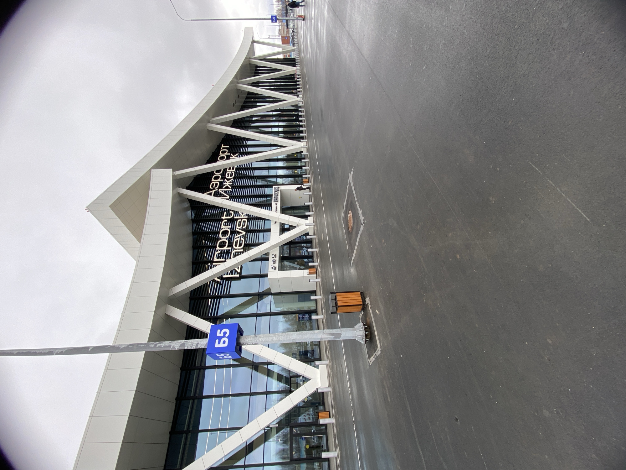 This image captures the modern terminal building of Izhevsk Airport in Izhevsk, Russia, on an overcast day. The airport building features a striking white, angular facade with a prominent roofline that forms a wave-like shape. Large, diagonal white beams create a zigzag pattern supporting the structure above a glass curtain wall that forms the main entrance.
Visible text on the building's facade reads "Аэропорт Ижевск" (Airport Izhevsk) in large black letters. Below this, a smaller sign indicates "Вход" (Entrance). In the foreground, a wide expanse of dark asphalt, likely a road or drop-off zone, appears wet, suggesting recent rain or snowmelt. White lane markings are visible on the pavement.
A tall metal light pole stands in the mid-ground, featuring a blue rectangular sign with "Б5" (B5) printed in white. Ice or frost appears to cling to the horizontal bar of this pole, indicating cool or cold weather conditions. Several wooden benches and black trash receptacles are placed along the sidewalk near the building. In the distance, a few blurred figures of people can be seen walking, and faint outlines of other buildings and trees are visible under the uniformly gray, cloudy sky. The scene depicts a functional airport entrance during daytime.