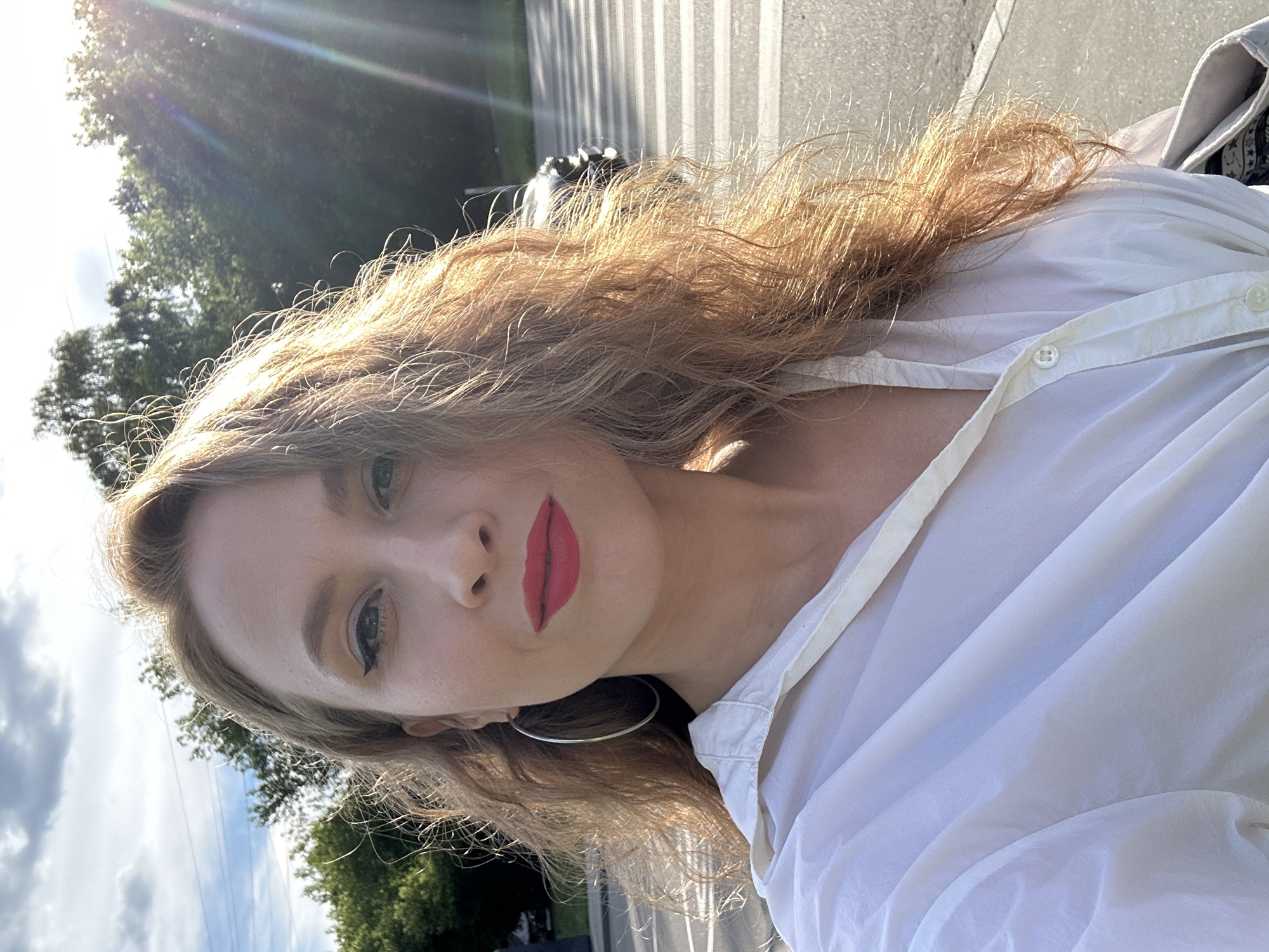 A vertical, close-up selfie captures a young woman outdoors. She is positioned in the lower right portion of the frame, with her head tilted towards the camera. The woman has long, wavy blonde hair that catches the sunlight, creating a halo effect. She is wearing a white collared shirt with visible buttons. Her makeup includes bold red lipstick and winged eyeliner. A large, silver hoop earring is visible in her left ear.
The background suggests an urban or suburban setting during the day. Trees and a cloudy sky are visible in the upper left and lower left of the image. A crosswalk is visible in the background, with a dark object, possibly a motorcycle, partially obscured behind the woman's head. The lighting indicates it is daytime, with strong sunlight creating lens flare in the upper left corner. The overall mood appears casual and relaxed. The image was taken in Moscow, Russia. No visible text is present in the media file.