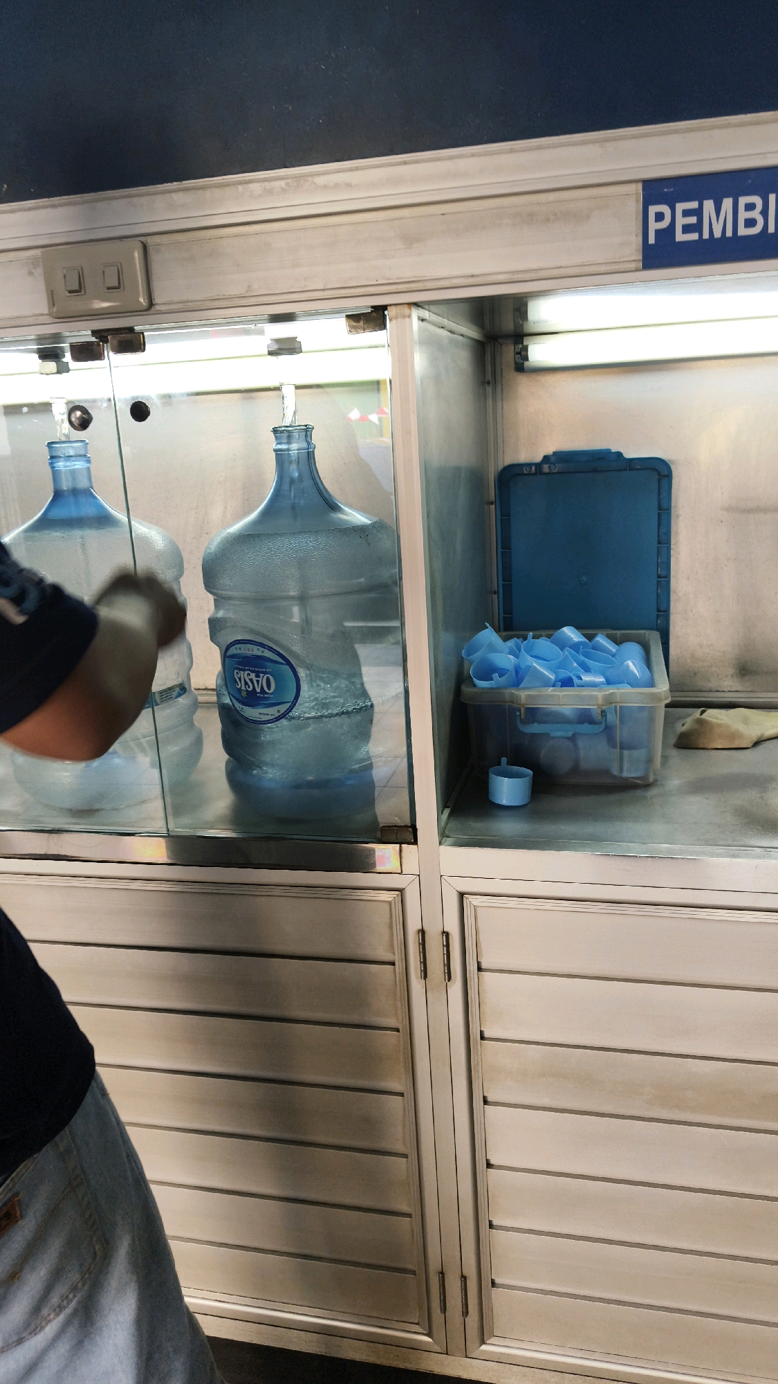 This image captures a close-up view of a water dispenser station, likely located in Bekasi, Indonesia, as suggested by the visible text "PEMBI". The scene is set within a glass-fronted cabinet with metal framing and white slatted doors at the bottom. Inside the cabinet, two large, clear plastic water jugs are prominently displayed. The jug on the right is full and has a label that reads "OASIS". A person's arm and shoulder, wearing a dark t-shirt and denim jeans, are visible in the lower left corner, suggesting they are interacting with the dispenser or one of the jugs.
To the right of the water jugs, a blue plastic container holds a stack of small, light blue disposable cups. Above this container, a blue lid is propped open, and a light fixture illuminates the interior of the cabinet. The cabinet itself appears to be made of brushed aluminum or a similar metal. A light switch with two toggles is mounted on the metal frame above the glass. The overall environment suggests a public space, possibly a convenience store or a dedicated water refilling station.