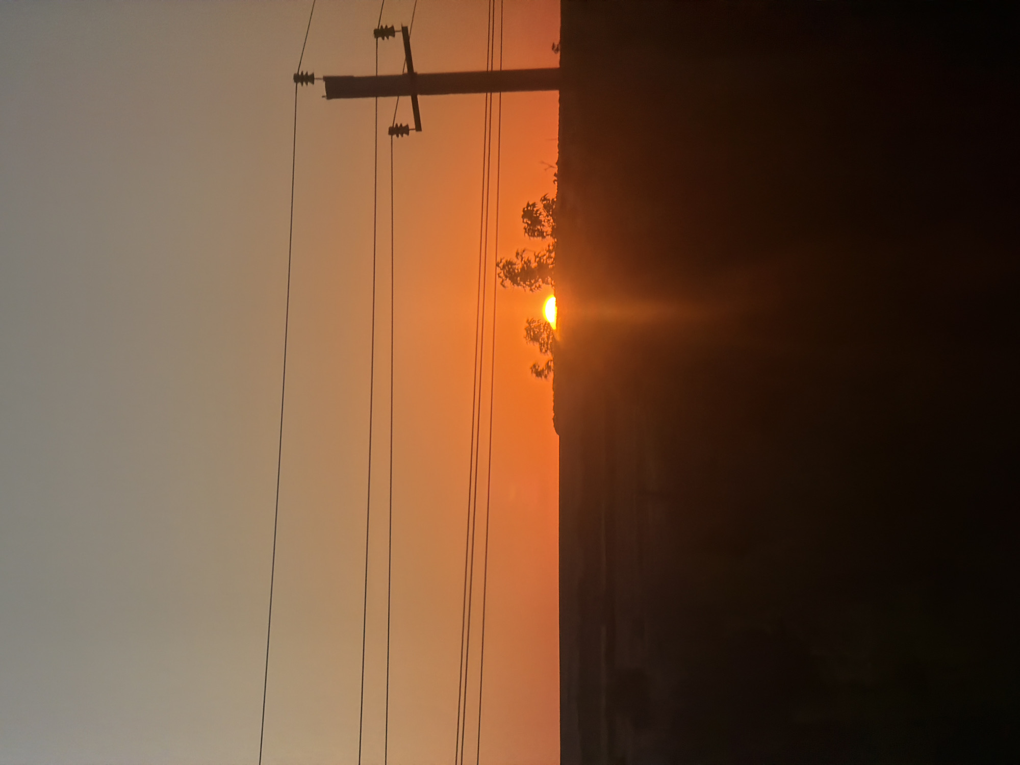 The image shows a sunset over a rural landscape, with a silhouette of trees and power lines against the orange sky. The sun is partially visible, just above the horizon, casting a warm glow. The foreground is dark, obscuring any details of the ground. The scene appears to be a tranquil evening in Karamysh, Russia.