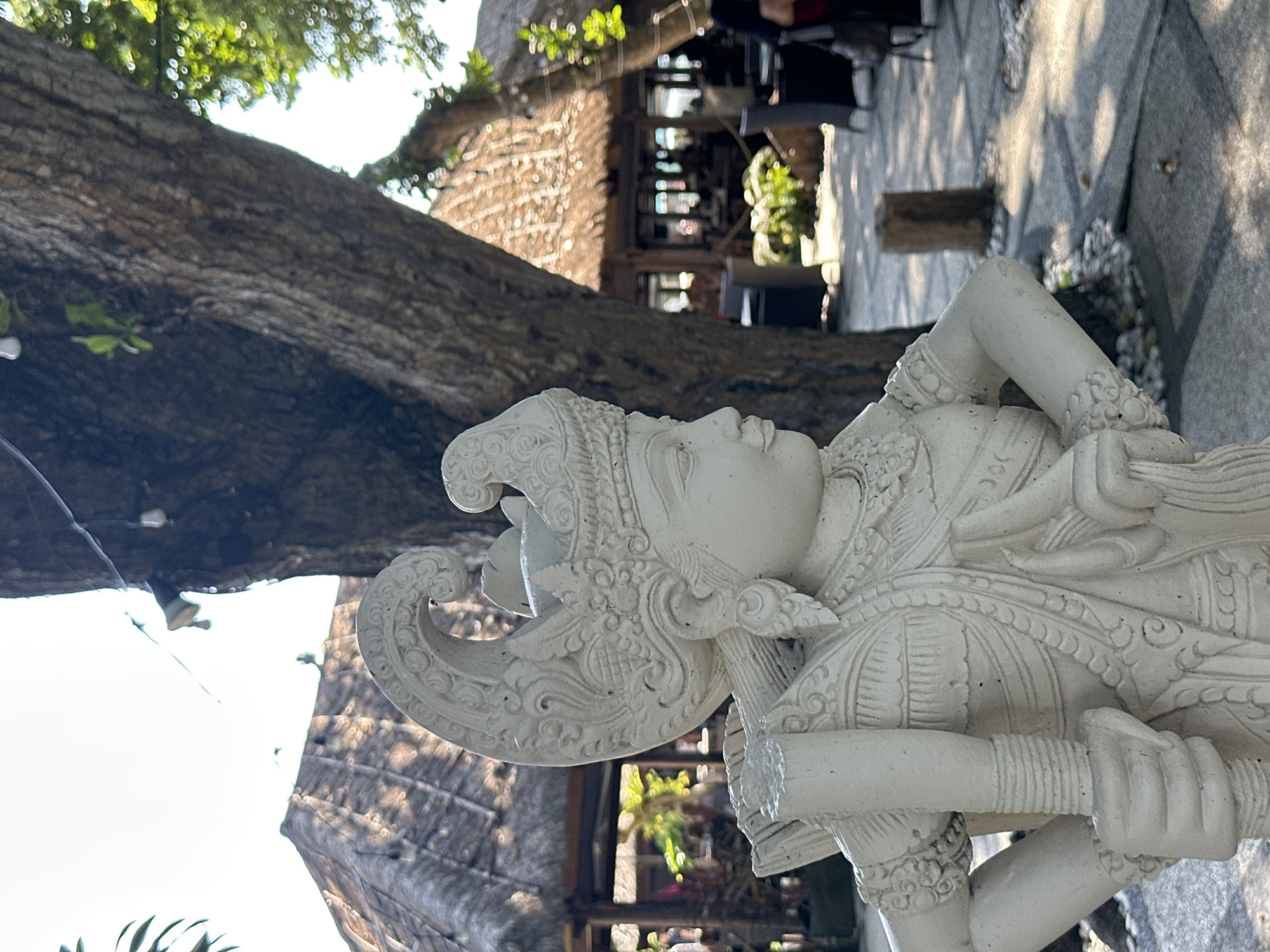 In Denpasar, Indonesia, a white stone statue of a Balinese figure stands prominently in the foreground, positioned in profile facing right. The statue, depicting a person adorned with a crown and traditional attire, holds an object in its left hand. The background reveals a thatched-roof structure, indicative of a restaurant or pavilion, with patrons seated at tables. A large tree trunk is visible behind the statue, and dappled sunlight filters through its leaves, casting shadows on the paved ground. The environment appears to be an outdoor seating area of a establishment.
