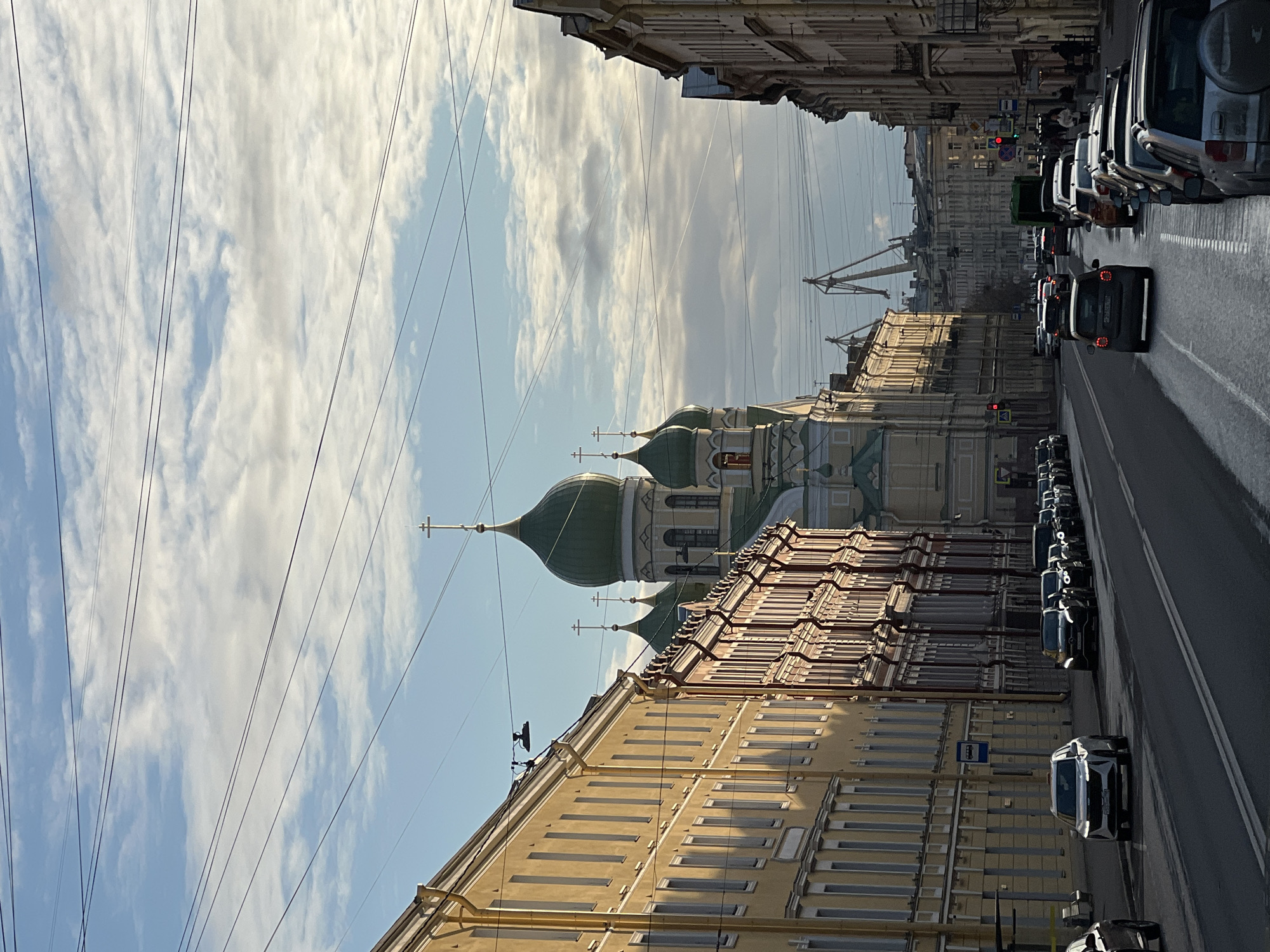 The image shows a street view in Saint Petersburg, Russia. A church with green domes and golden crosses stands out amidst the surrounding buildings. A row of buildings lines the street, with a color palette of light yellow and beige. A road with parked cars is visible on the right side of the frame. Overhead power lines stretch across the sky, which is partially covered by clouds. The overall scene is bathed in sunlight, creating shadows on the buildings and street.