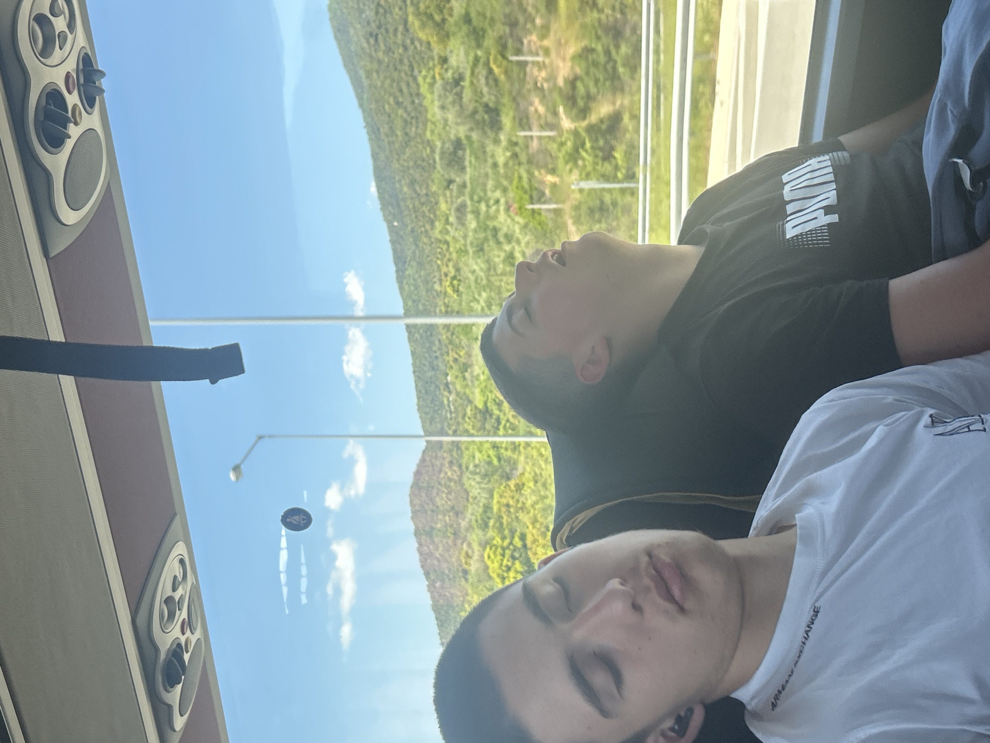 Two people are seated on a bus. One wears a black Puma shirt and the other a white Armani Exchange shirt. Both are asleep. The bus travels along a road, with a view of forested mountains visible outside the window. A street lamp and a sign with the Greek letter omega are visible through the window as well. The vehicle appears to be traveling through Vrasna, Greece. The interior of the bus is visible with AC and speaker.