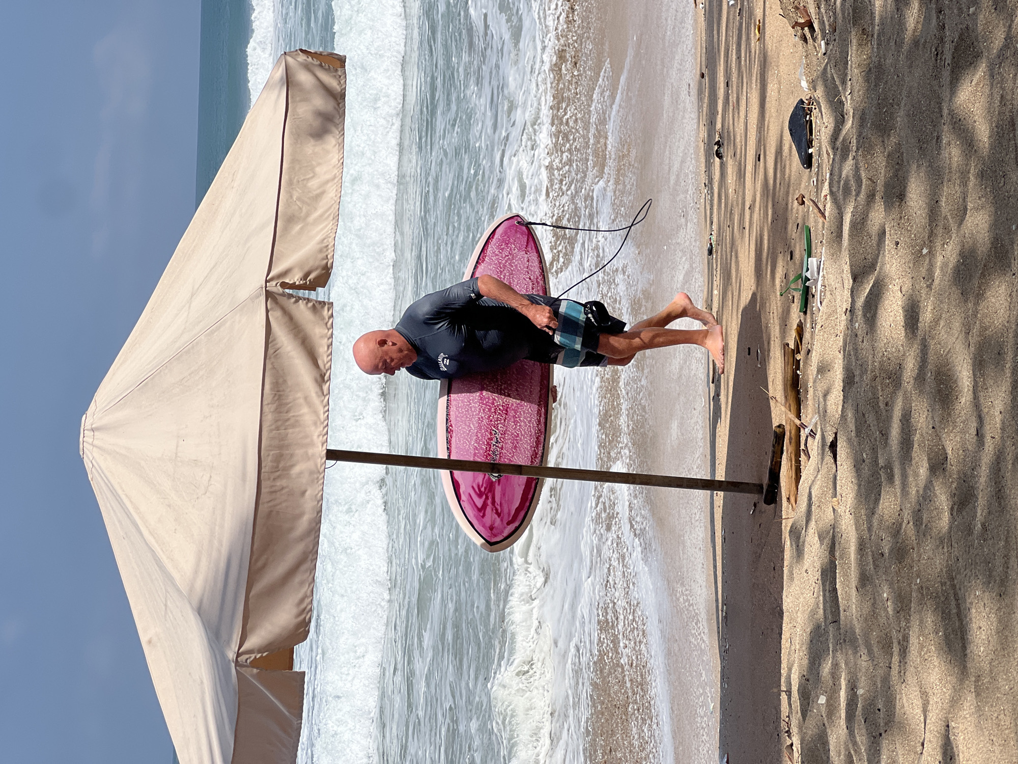 A bald, fair-skinned man in a dark rashguard and checkered swim trunks walks on a sandy beach in Kuta, Indonesia, holding a pink surfboard. He is walking from left to right under a large, light-colored beach umbrella, with the ocean and waves in the background. The surf is moderate. The sand is littered with debris including driftwood and plastic.