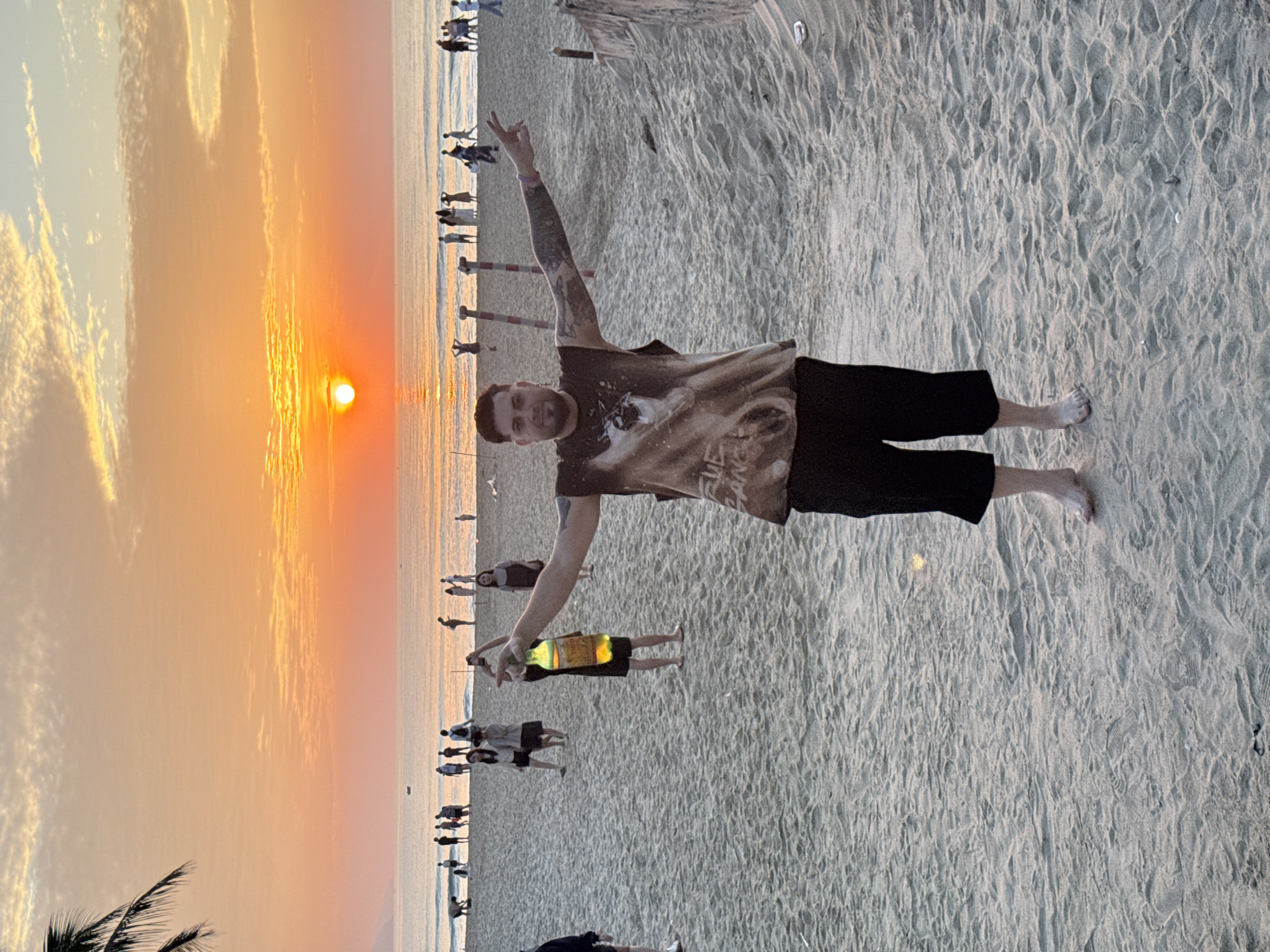 A man stands on a sandy beach at sunset in Da Nang, Vietnam, with his arms outstretched. He is wearing a dark t-shirt with a graphic print and black shorts. He is barefoot. In the background, other people are visible on the beach and in the shallow water. The sun is setting over the ocean, casting an orange glow across the sky and water. The sand is light brown and appears to be undisturbed except for footprints. The scene is peaceful and serene.