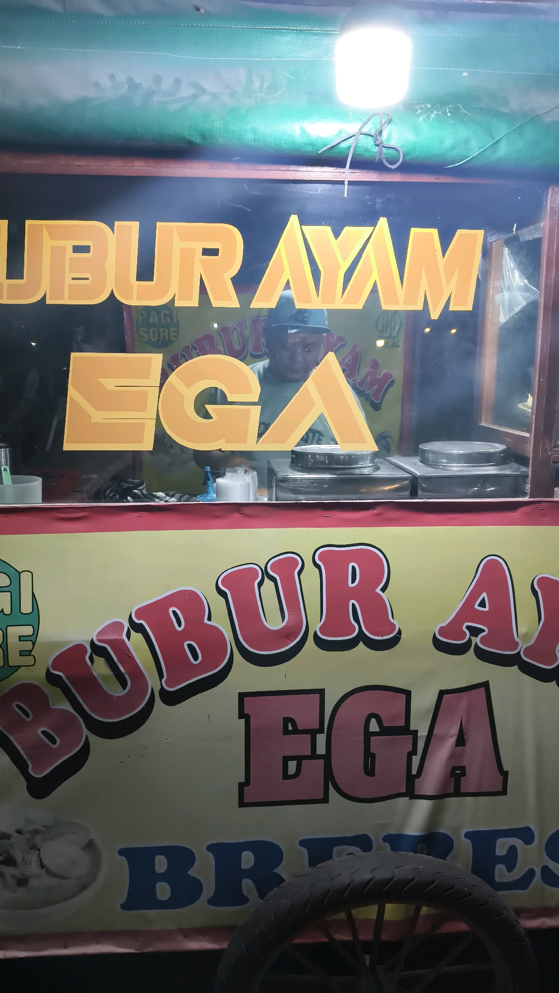 A food cart with the words "BUBUR AYAM EGA" (Chicken porridge EGA) is displayed prominently. The words are written in large, bold letters, with "BUBUR AYAM" in yellow and "EGA" in orange. The cart also has "PAGI SORE" (Morning Afternoon) written on it, indicating it is open all day. A person, wearing a cap and a green shirt, is visible behind the counter, possibly preparing food. The cart is illuminated by a bright overhead light. The scene is set at night, with the cart appearing to be on a street in Satriajaya, Indonesia, as indicated by the text on the cart. The bottom of the image shows a wheel of the cart and the words "BR..." which may be part of "BROTO" or another word.
