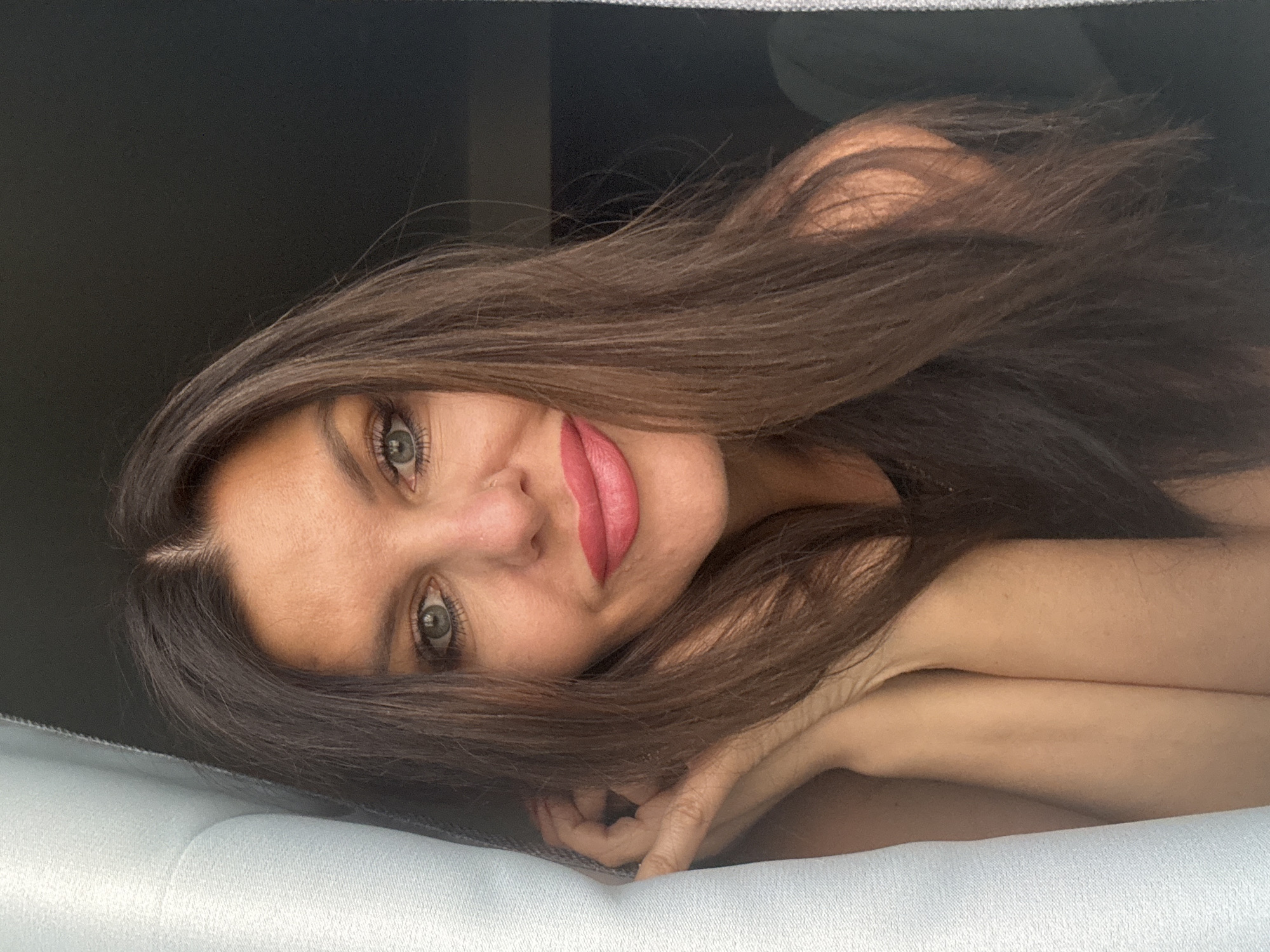 This is a close-up, horizontally oriented photograph of a woman lying on a light-colored surface, possibly a bed or couch. She is positioned on her side, with her head tilted and her hair falling around her face. The woman has long, dark brown hair, and her eyes are a striking green. She is wearing bright pink lipstick, which stands out against her fair skin. Her left hand is visible near her face, resting on the surface. The background is dark and appears to be a solid surface, possibly a wall or furniture. The lighting suggests an indoor setting, and the overall mood is intimate and relaxed. The photograph is taken from a slightly elevated angle, looking down at the woman. There is no visible text in the image.