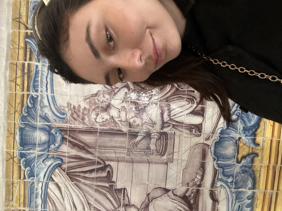 An angled selfie captures a young adult female positioned in the foreground, smiling faintly towards the camera. She presents with dark brown hair, dark eyes, and fair skin, wearing a black high-neck garment and a rose gold chain draped over her right shoulder. The background features a large, ornate *azulejo* (tile) panel, identifiable as being from the Palácio dos Marqueses de Fronteira in Lisbon, Portugal.
The *azulejo* panel depicts a historical or biblical narrative rendered predominantly in sepia (brown/purple wash) tones on a white tile background, framed by blue and white decorative borders and a yellow-gold patterned strip at the top and bottom. The scene within the panel includes several robed figures: a central figure kneels with outstretched hands in an apparent gesture of supplication, directed towards a partially visible figure wearing sandals on the far left. To the right of the kneeling figure, a standing woman with crossed arms and two armed figures, possibly soldiers wearing helmets and holding spears, are depicted observing the scene. An architectural element resembling a fluted column is visible to the left of the kneeling figure. The tiles display visible grout lines and fine craquelure, indicative of their age.