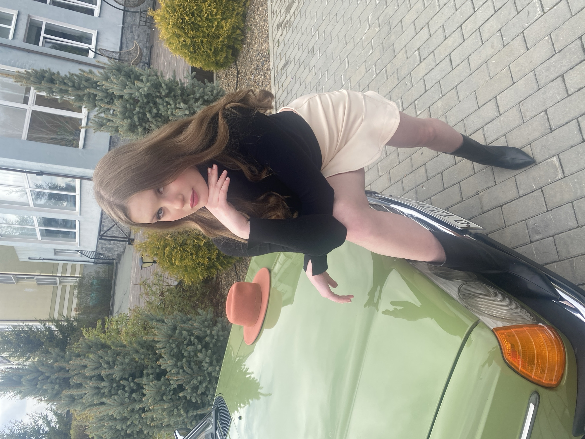 A young woman with long, light brown hair is reclining on the hood of a light green car. She is wearing a black long-sleeved top and a light-colored skirt, and black boots. Her right hand is near her face, and her left hand is extended toward the car's hood. A brown hat sits on the hood of the car. The car's front end is visible, including the headlight and bumper.
The background includes a brick walkway, buildings, and evergreen trees. The location is likely Yekaterinburg, Russia.