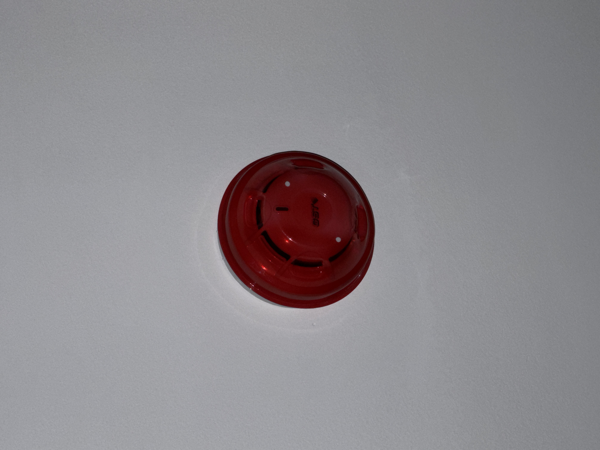 A close-up, slightly angled shot shows a red fire alarm device mounted on a white ceiling. The device is circular with several vents and a small, circular indicator light. The brand name "GST" is visible in white lettering on the device's surface. The ceiling appears to be smooth and textured. The lighting suggests an indoor setting. There are no people or other objects visible in the image.