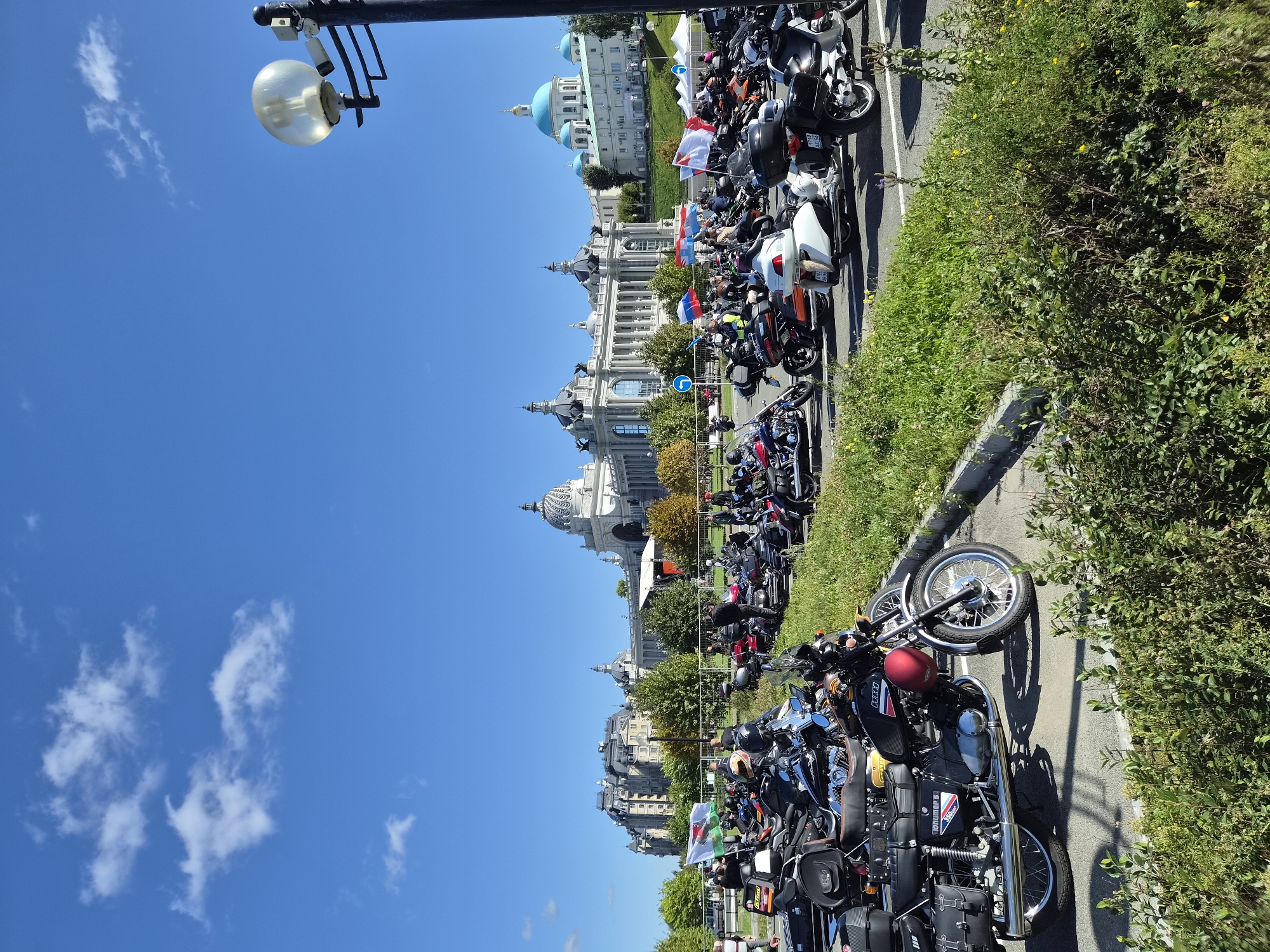 In Kazan, Russia, a large gathering of motorcycles is parked in front of a grand architectural complex on a clear, sunny day. The complex features buildings with domes and classical architectural elements, possibly a palace or government building, and a church with blue domes. A streetlamp with multiple bulbs and two security cameras is visible in the foreground, to the right. Numerous people, primarily men, are seen standing or interacting around the parked motorcycles, suggesting a motorcycle rally or event. The motorcycles are of various makes and models, including cruisers and touring bikes, with some adorned with flags, such as the Welsh flag. The environment includes a paved parking area, lush green grass, and trees with autumn foliage. The sky is a vibrant blue with scattered white clouds.