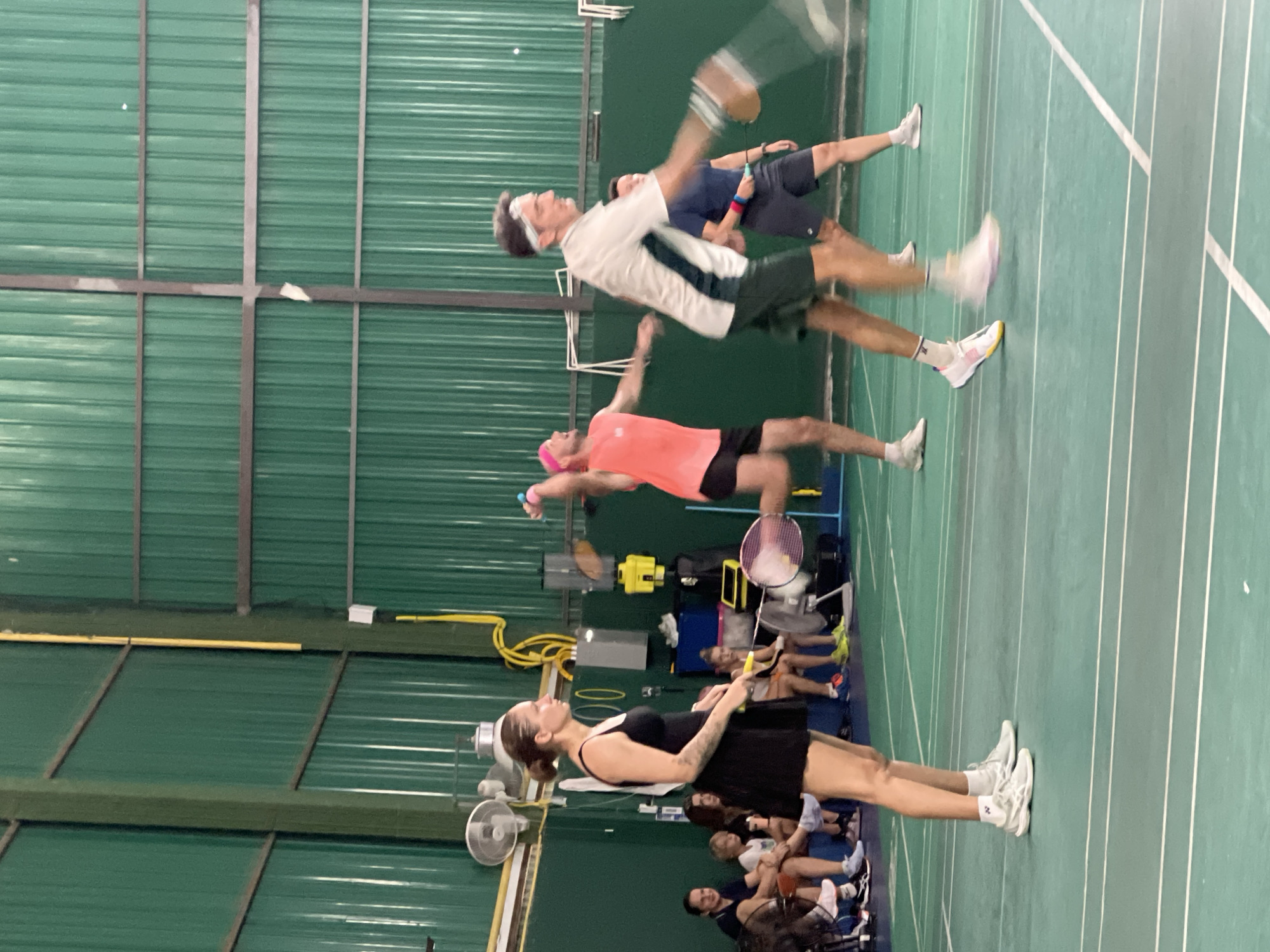 In a badminton court in Ban Rak, Thailand, a co-ed doubles match is underway. The court is indoors with a green, corrugated metal wall serving as the backdrop. A woman in a black dress and athletic shoes stands on the left side of the court, holding a badminton racket. To her right, a man in a white and green shirt, dark shorts, and a headband is in motion, mid-swing with his racket. Further to the right, another man in a coral-colored tank top and dark shorts is also captured in motion, his arm extended as if playing. In the background, spectators are seated, and various sporting equipment is visible. The floor of the court is a vibrant green with white lines marking the boundaries.