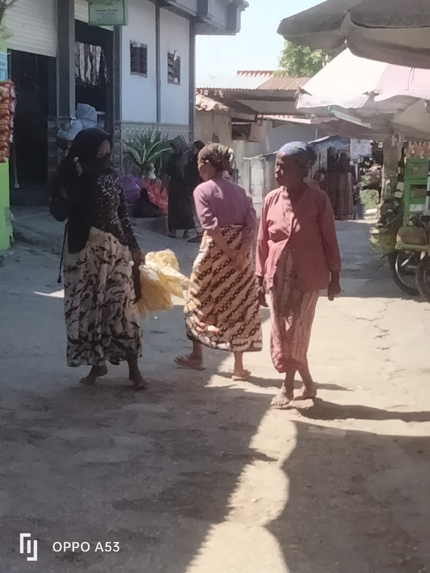 Three women walk along a dusty street in what appears to be a market area, likely in Badung, Indonesia, given the camera's watermark. The woman on the left is wearing a dark patterned headscarf and a long, light-colored skirt with a dark abstract pattern. She is carrying a bundle of light-yellow material. The middle woman wears a purple top and a long skirt with a diagonal zigzag pattern in beige and brown. The woman on the right has a blue headscarf and is wearing a pink long-sleeved top and a striped skirt in pink and white. The street is unpaved and there are buildings and market stalls visible in the background, including a green structure with a wheel, possibly part of a cart or motorcycle. The scene is lit by strong sunlight, casting distinct shadows on the ground.