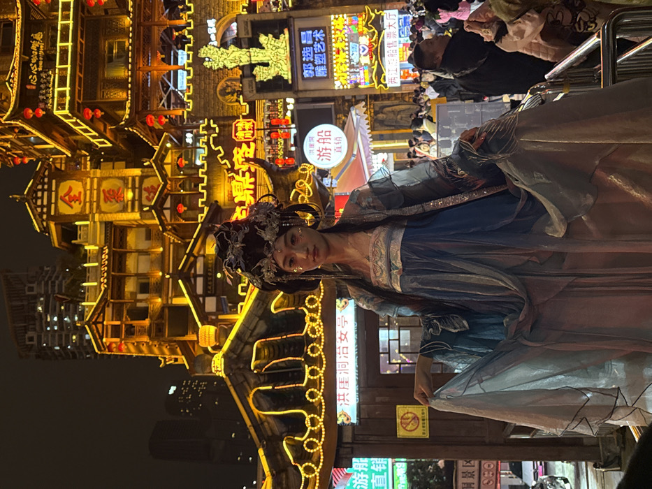 A woman in an elaborate blue and silver gown with matching headpiece stands in front of a brightly lit, ornate building at night. Chinese characters and illuminated signs surround her, creating a vibrant and busy atmosphere. The woman appears to be posing, and the overall impression is one of cultural performance or costume play in a lively urban setting.
