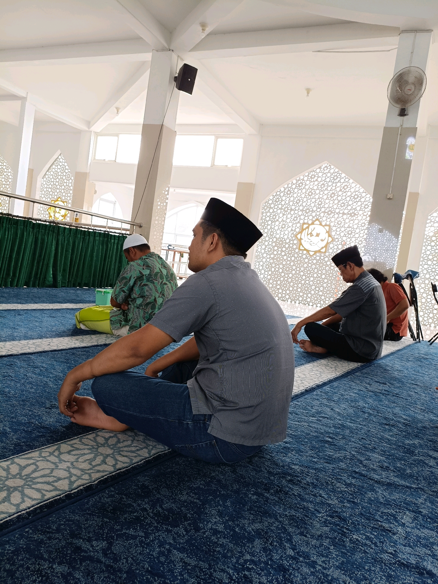 In the city of Maguwoharjo, Indonesia, inside a mosque, a group of men are seated on a blue carpeted floor, facing a wall adorned with intricate white geometric patterns. The men are positioned on prayer mats, some in a seated position with legs crossed, others in a more upright posture. One man in the foreground wears a grey striped shirt and blue jeans, his back to the camera. To his left, a man in a green patterned garment and a white cap is also seated. Further in the background, another man in a grey shirt and black pants is seen. The mosque's interior features high ceilings with white beams and illuminated by natural light filtering through windows. Industrial fans are mounted on the walls, and speakers are visible on the ceiling. A green curtain hangs along one wall, and a yellow cushion is placed on the floor. The scene captures a moment of quiet contemplation or prayer within the religious space.
