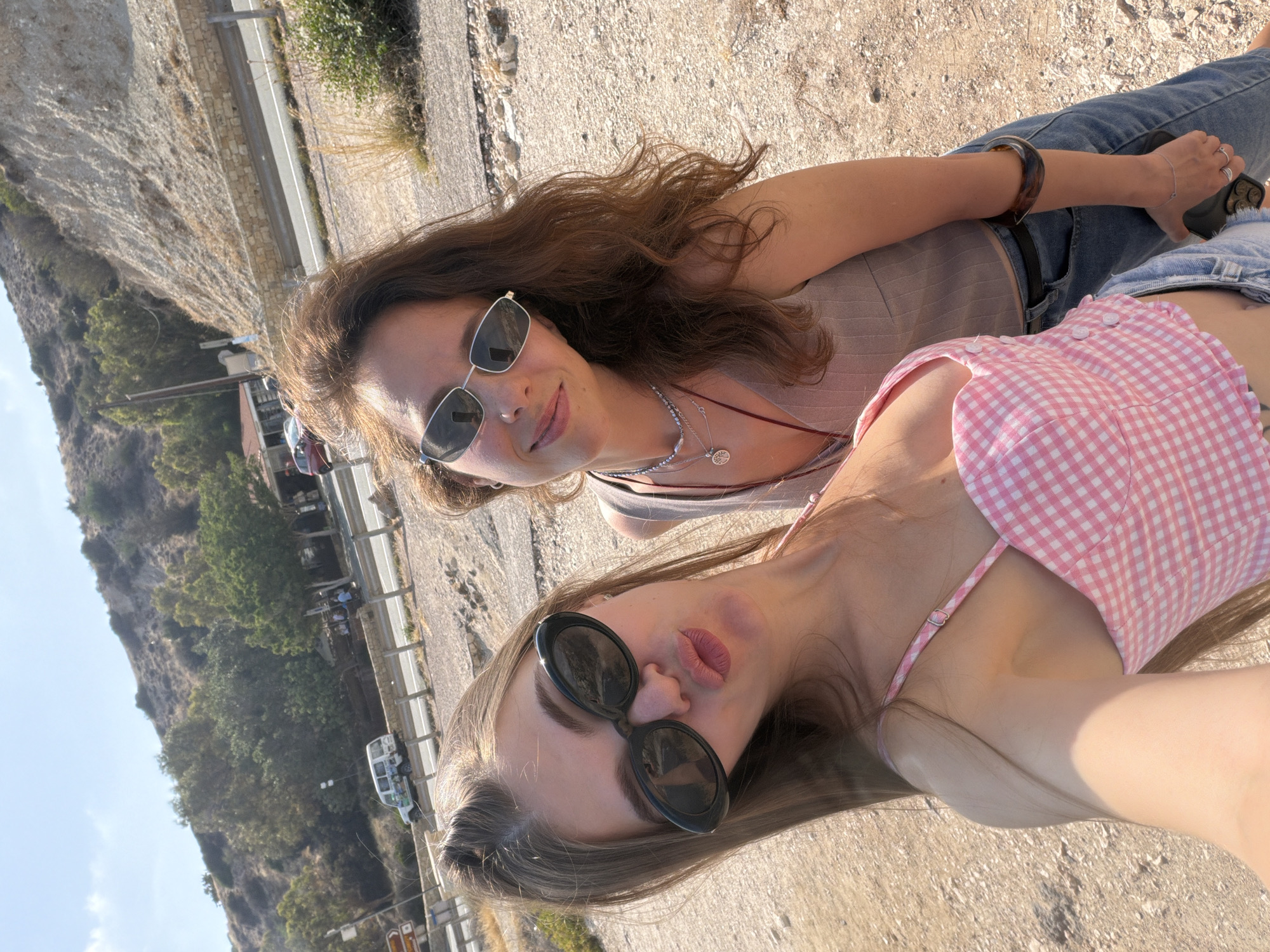 The image features two young women outdoors, possibly on a rocky or gravelly hillside, with a road and vegetation visible in the background. The scene appears to be a sunny day, suggesting it is daytime.
The woman in the foreground is wearing a pink and white checkered top and black sunglasses. She is looking towards the camera with her lips slightly parted. The woman behind her is wearing a gray top and silver sunglasses, and she is smiling towards the camera. Both women are wearing casual clothing.
The background shows a steep, rocky incline on the left and a road with a white guardrail on the right. Trees and bushes are growing on the hillside. There are some vehicles visible on the road, including a white van. The ground in the foreground is covered in small rocks and pebbles.
The overall mood of the image is relaxed and cheerful, suggesting a casual outing or vacation. The lighting indicates a bright, sunny day. No visible text is present in the image.