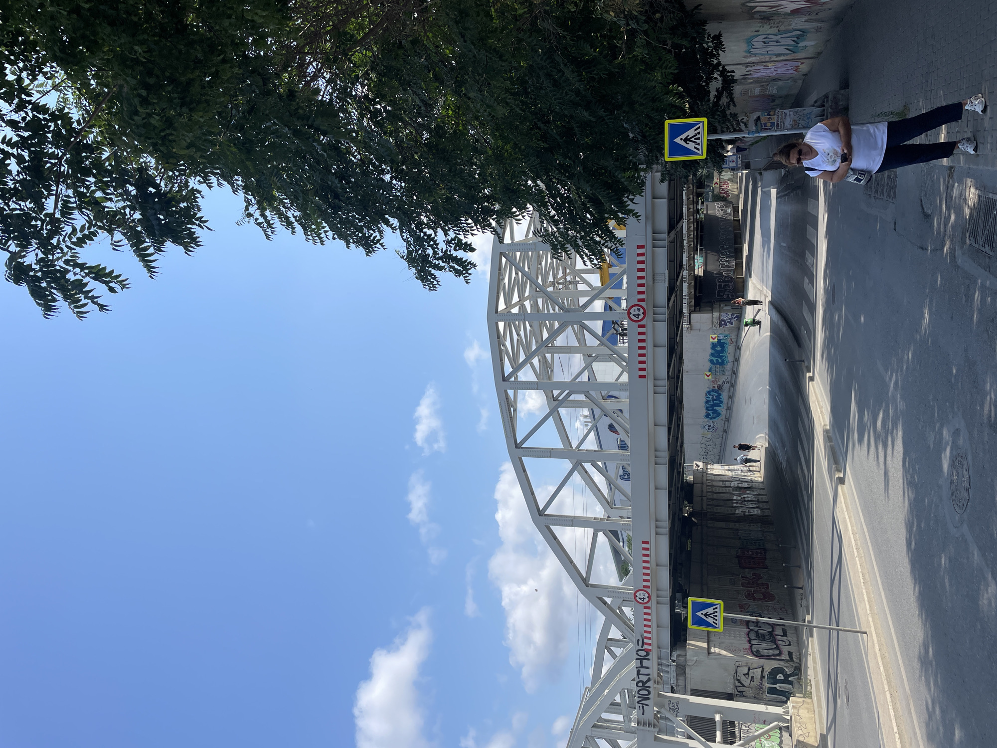 The image depicts a street scene under a white metal truss bridge, likely in Kadıköy, Turkey, given the context. The bridge has graffiti on its support pillars. A woman in a white t-shirt and dark pants is standing on the sidewalk, holding a phone. To her right, a yellow pedestrian crossing sign is visible. The road surface is asphalt with lane markings. Further down the road, under the bridge, two individuals are walking. Another pedestrian crossing is marked on the road. The sky is blue with scattered white clouds. A large tree with green leaves is on the right side of the image, partially obscuring the view. A red circular sign with "4.40" is visible on the bridge.