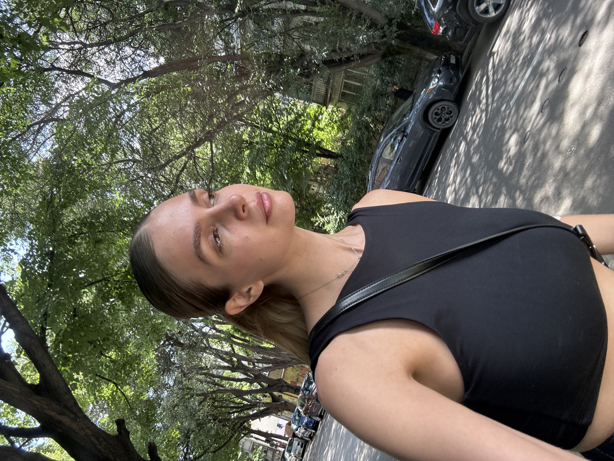 A young woman with blonde hair pulled back in a ponytail and fair skin is walking on a sun-dappled street lined with trees. She is wearing a black, sleeveless crop top and a thin silver necklace. Her right shoulder and arm are visible in the foreground. In the background, cars are parked along the side of the road, and buildings with balconies are visible through the foliage. The scene appears to be in an urban environment, likely Tbilisi, Georgia, given the architecture and tree-lined streets typical of the city. The lighting suggests it is daytime, with sunlight filtering through the leaves, creating patterns of light and shadow on the pavement.