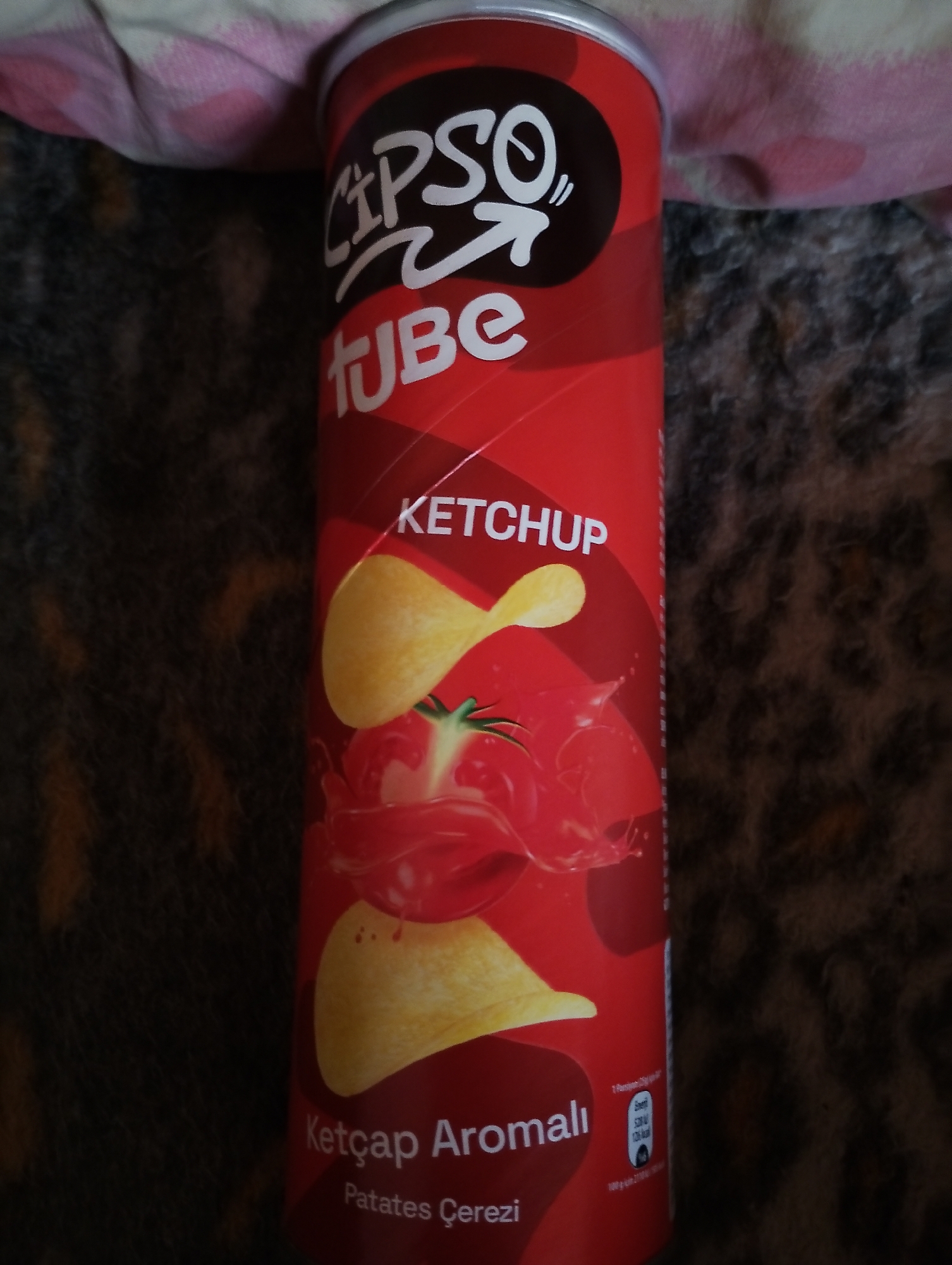 The image shows a close-up, vertical view of a red cylindrical tube of "ÇipsOTUBE" brand potato chips. The logo "ÇIPSOTUBE" is prominently displayed at the top in white lettering. Below the logo, the flavor "KETCHUP" is written in white capital letters. A graphic of a tomato and several potato chips are depicted on the tube. Towards the bottom, the text "Ketçap Aromalı Patates Çerezi" is visible, indicating the flavor is ketchup-flavored potato chips. The tube is placed on a dark, textured surface that appears to be fabric, possibly a blanket or rug. A corner of a pink and white checkered fabric item is visible in the upper left of the frame. The lighting is somewhat dim, with a soft focus that suggests the image was taken indoors. There are no people or identifiable geographical locations present in the image.