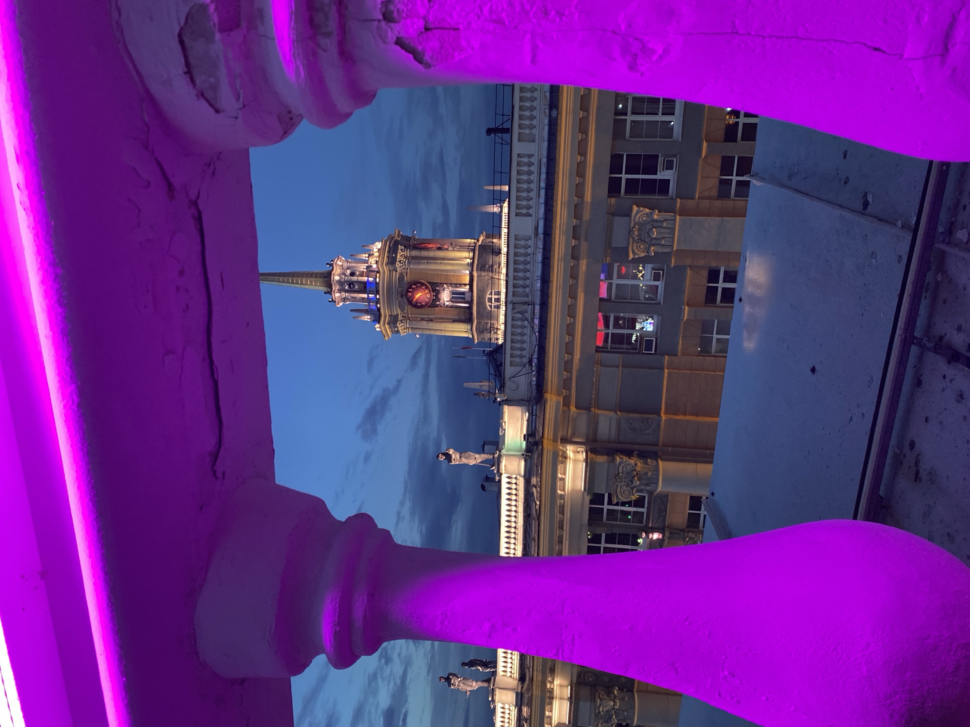 The image captures a nighttime or late twilight urban scene, observed from an elevated vantage point behind a brightly illuminated foreground structure. Two substantial, sculpted concrete balusters, one on the left and one on the right, frame the view, connected by a horizontal rail. These concrete elements are intensely bathed in artificial magenta-purple light, highlighting their rough texture and minor surface cracks.
Beyond the balustrade, a multi-story building with classical architectural features occupies the midground. Its roofline is adorned with a decorative balustrade, and at least three dark, static figures, identifiable as statues, stand along the roof: two on the left and one on the right. The building's facade is illuminated with warm light, revealing multiple rectangular windows with white frames; some windows show interior light. Ornate architectural details, possibly corbels or capitals, are visible between window sections on the upper stories.
In the background, against a dark blue twilight sky with scattered clouds, stands the distinctive, illuminated clock tower of the Yekaterinburg City Administration Building, located at Lenin Avenue, 24, Yekaterinburg, Russia. The tower is brightly lit with warm yellow and orange light, accentuating its ornate multi-tiered structure and a tall, slender spire. At least two prominent clock faces are visible, illuminated with warm light and displaying red markings or hands. Minor accents of blue light are present near the upper sections of the tower's base. No active human figures are discernable within the visible areas of the scene.