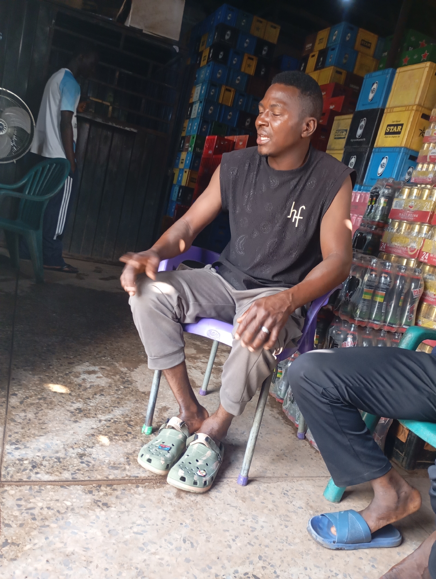 The image depicts an outdoor scene, likely a store, in Shendam, Nigeria. In the foreground, a man is seated on a purple plastic chair. He is wearing a sleeveless black shirt with a white "hf" logo and grey pants. His right hand is resting on his knee, and he appears to be speaking, with his mouth open. On his left hand, a ring is visible. He is wearing green clogs. To the right, another person is partially visible, wearing black pants and blue flip-flops. Behind the seated man, stacks of colorful crates, likely containing beverages, are piled high. These crates are predominantly blue, yellow, and red, with some bearing labels such as "STAR" and "NB". In the background, another person, wearing a white t-shirt with blue accents and black pants with white stripes, stands near a metal fan and a dark wooden structure. The ground is a speckled concrete surface, with some patches of moisture or staining. The lighting suggests it is daytime, with shadows cast on the ground.