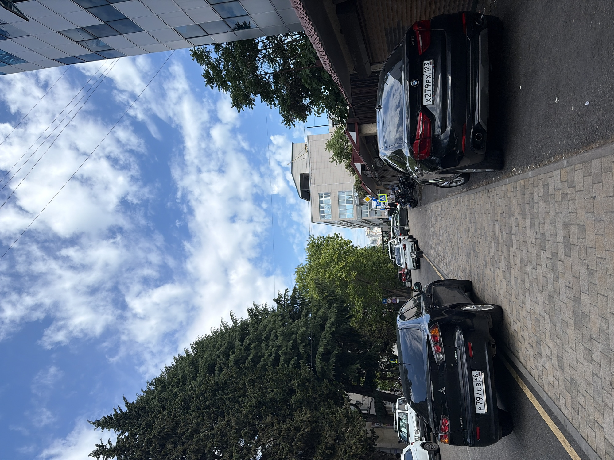 This image captures a street scene in Sochi, Russia, under a partially cloudy blue sky. In the foreground, a black Mitsubishi Lancer sedan is parked on a cobblestone sidewalk, facing away from the viewer. Its license plate reads "P797CB 46 RUS". To the right, a black BMW X6 is also parked on the street, facing the same direction. Its license plate displays "X279PX 123 RUS". Several other vehicles, including a white SUV, are visible further down the street. Tall evergreen trees line the left side of the road, casting shadows on the sidewalk. A modern, multi-story building with a reflective facade stands on the right, partially obscuring a row of shops with brown awnings. The street appears to be a busy urban area, with utility lines crossing overhead.