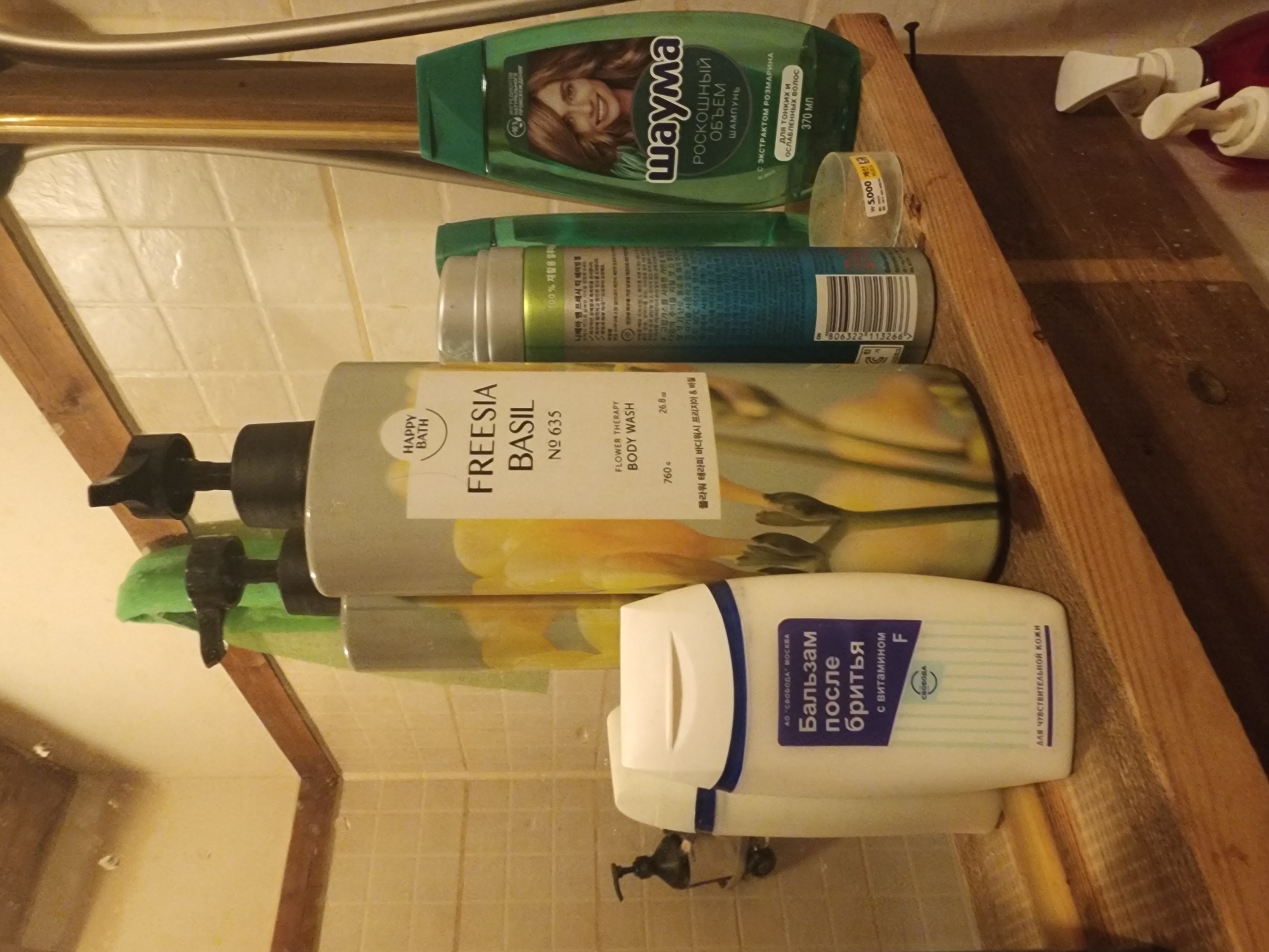 The image shows a cluttered bathroom shelf, possibly in a residential setting in Pyeongtaek-si, South Korea. The shelf is made of rough, unfinished wood and is mounted on a tiled wall. Various personal care products are displayed, including a green bottle of shampoo with a picture of a woman, a tall, light-colored bottle with a label that says "FREESIA BASIL," and a white bottle with Russian text that appears to be after-shave balm. There are also other bottles and containers, some partially visible. The overall impression is one of a well-used and somewhat unorganized bathroom.