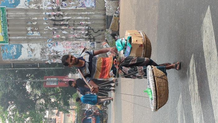 An urban street scene in Dhaka, Bangladesh is depicted in daylight. A man, approximately 30-40 years old, with dark hair and a beard, walks from right to left across an asphalt road, his gaze directed slightly downward and forward. He wears a horizontal striped short-sleeved shirt (grey, white, orange, blue segments) and a dark patterned lungi, tied at the waist. He wears dark sandals. He carries a wooden or bamboo pole across his right shoulder, acting as a yoke, from which two large, round, woven baskets are suspended by ropes. The left basket, in the foreground, contains light-colored material, with a small green plastic bag placed on top. The right basket holds a cardboard box, upon which rests a blue and green digital weighing scale.
In the background, on the left, several other individuals are visible on a sidewalk, including a man in a blue shirt and another in a black t-shirt. A blue rickshaw is partially visible on the far left, along with a dark two-wheeled vehicle and other indistinct figures. The road surface is asphalt, with faded white zebra crossing markings visible in the lower foreground. A concrete curb separates the road from a sidewalk. A large concrete pillar or wall occupies the right side of the background, heavily plastered with multiple layers of torn paper posters and graffiti, featuring Bengali script and various colors. Trees with green foliage are visible further in the background on the left, along with a slender dark street lamp pole and a red rectangular signpost. Scattered debris is present near the base of the pillar.