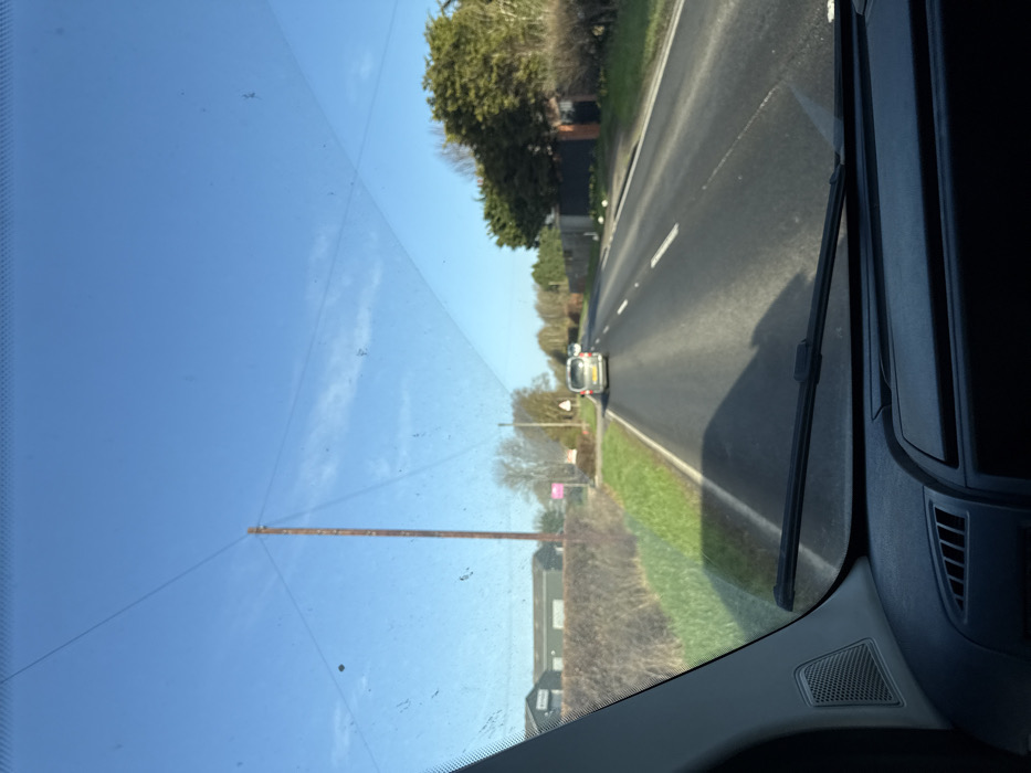 The image depicts a forensic view from the interior of a vehicle, facing forward on an asphalt road under a clear, bright blue sky with sparse, wispy clouds. This scene is located on an unspecified rural road within the Borough of Rushcliffe, United Kingdom.
In the immediate foreground, the top of a vehicle's dashboard is visible, featuring a speaker grille on the left and a portion of the windshield wiper blade at the bottom center. The windshield is visible, displaying accumulated dirt/dust and a faint pattern of cracks or scratches.
The road ahead is a single-carriageway with two lanes, marked by white dashed lines. A silver or light-colored minivan or MPV is observed in the distance, traveling away from the viewer, with another less distinct vehicle further behind it. Shadows cast by the vehicles are visible on the road surface, indicating sunny conditions.
To the left of the road, there is a grassy verge leading to a dense, trimmed hedge. Behind the hedge stands a tall, dark-colored utility pole with multiple overhead wires extending across the sky. Further back and to the left, several large, dark grey industrial or agricultural buildings, possibly barns or sheds, are present. Near a small track or entrance on the left, a distinct pink sign, likely a real estate advertisement, is visible alongside a white triangular road sign with a red border and an 'X' symbol, indicating a crossroads or junction ahead.
To the right of the road, a similar grassy verge abuts a continuous line of dense trees and hedges, which partially obscure any structures beyond them. No people are discernible within the frame.