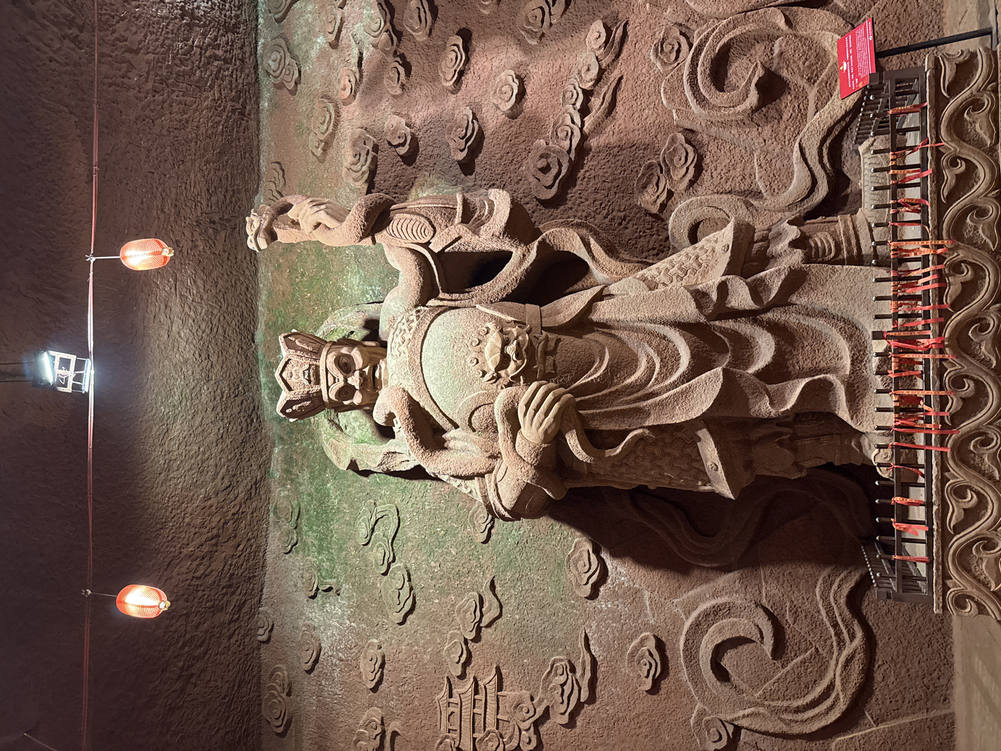 The image shows a stone carving of a figure in an unknown city. The figure wears a crown and robe, and is surrounded by carved clouds and other decorative elements. There is a metal rack with red ribbons tied to it next to the figure. The lighting is dim, with two red lanterns and a set of square lights providing illumination. The walls appear to be made of rough-hewn stone.
