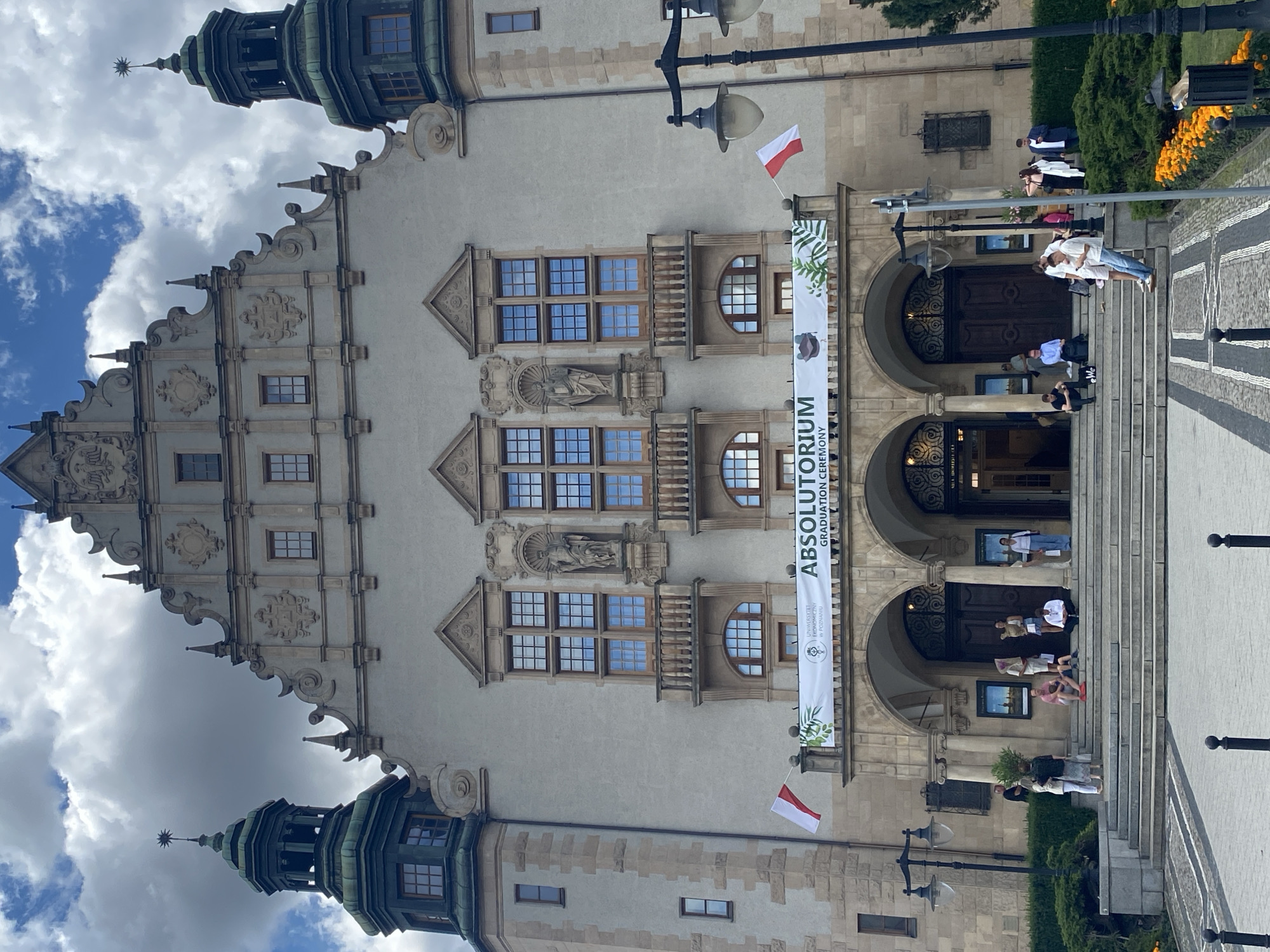 The image depicts the facade of the University of Economics in Poznan, Poland, with a clear sky and dramatic clouds. The grand building features two prominent towers with green roofs, ornate architectural details, and multiple windows. A banner above the entrance reads "ABSOLUTORIUM GRADUATION CEREMONY" and displays the university's logo. Two Polish flags are visible on either side of the banner. A group of people are gathered on the wide stone steps leading up to the arched entrance, some sitting and some standing, engaged in conversation or looking at their phones. The surrounding area includes decorative bushes and flowerbeds with orange flowers. A lamppost stands to the right of the steps.